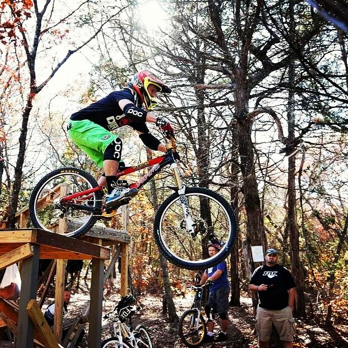 A mountain biker in a green and black outfit is mid-air, jumping off a wooden ramp in a forested area. Sunlight filters through the trees, and there are a few spectators watching nearby. The rider is wearing a helmet and protective gear, showcasing an action-packed moment in mountain biking. Big Cedar Wilderness Trails mountain bike trail.