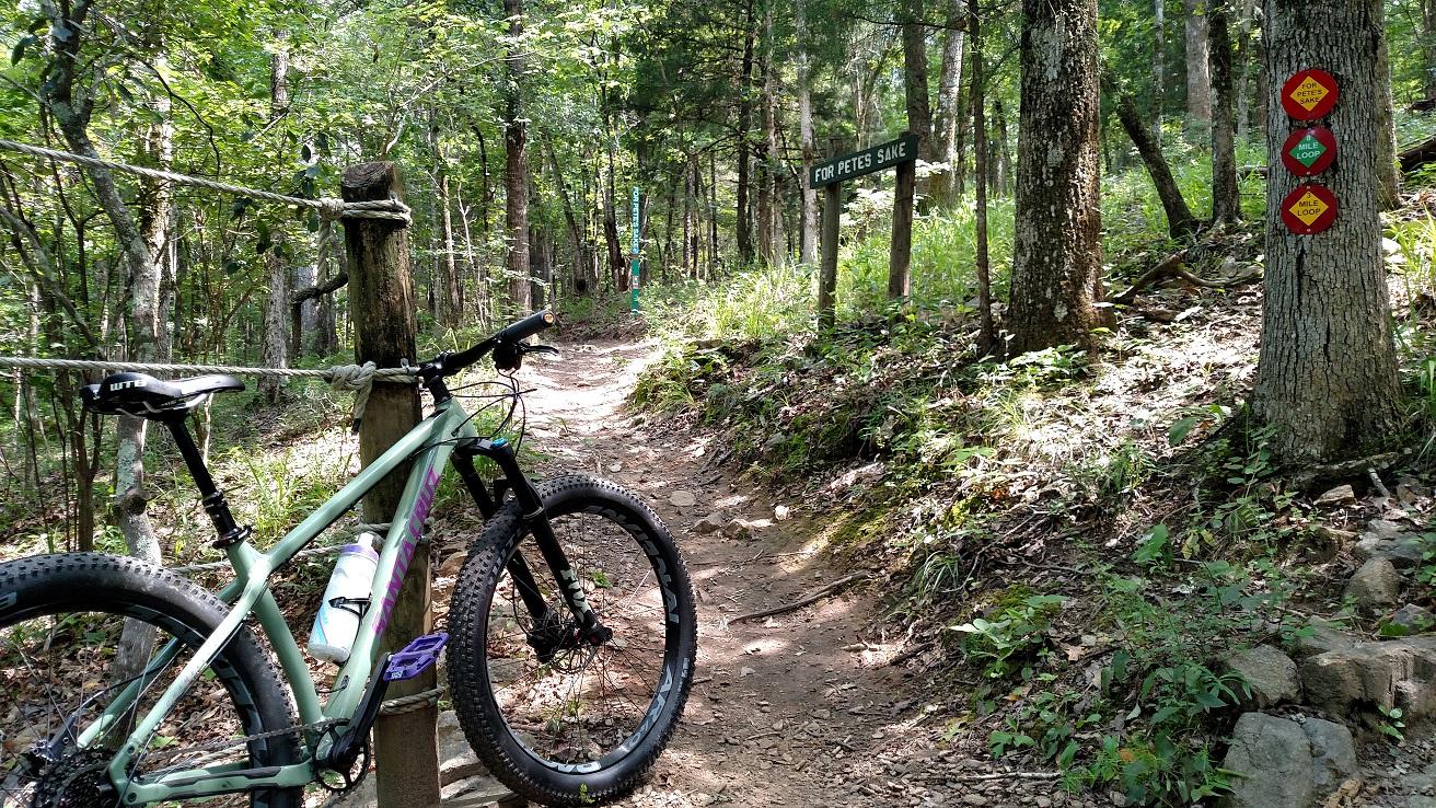 Santa Cruz Chameleon: A mountain bike leans against a wooden post at the entrance of a wooded trail, with a sign reading "For Pete's Sake" in the background. The path is lined with trees and greenery, indicating a natural outdoor setting, and there are trail markers visible nearby.