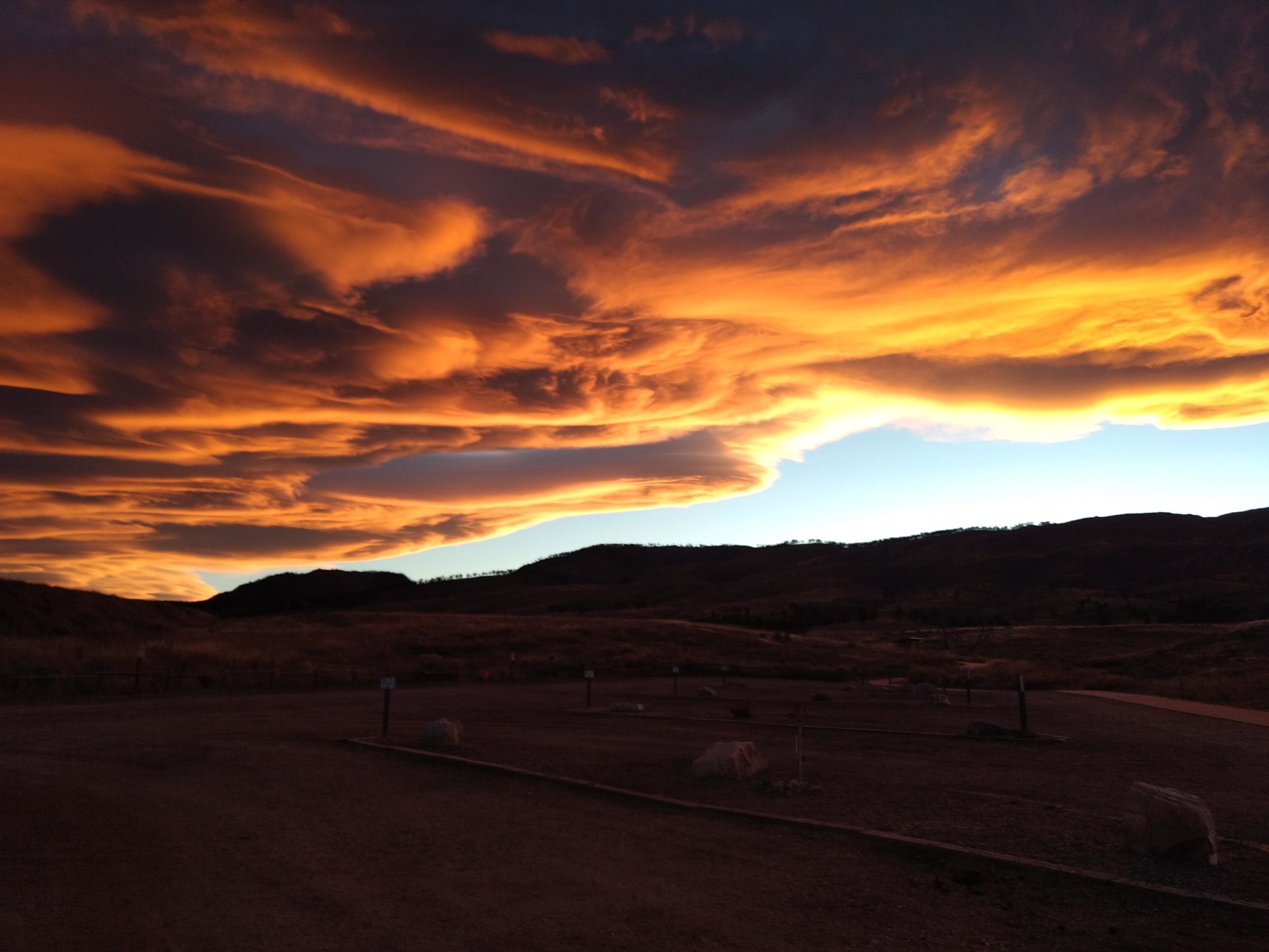 A dramatic sunset featuring vibrant orange and yellow clouds swirling above a mountainous landscape, casting a warm glow over the horizon. The scene captures the transition from day to night with a darkening sky and silhouettes of hills in the background. Bobcat Ridge mountain bike trail.