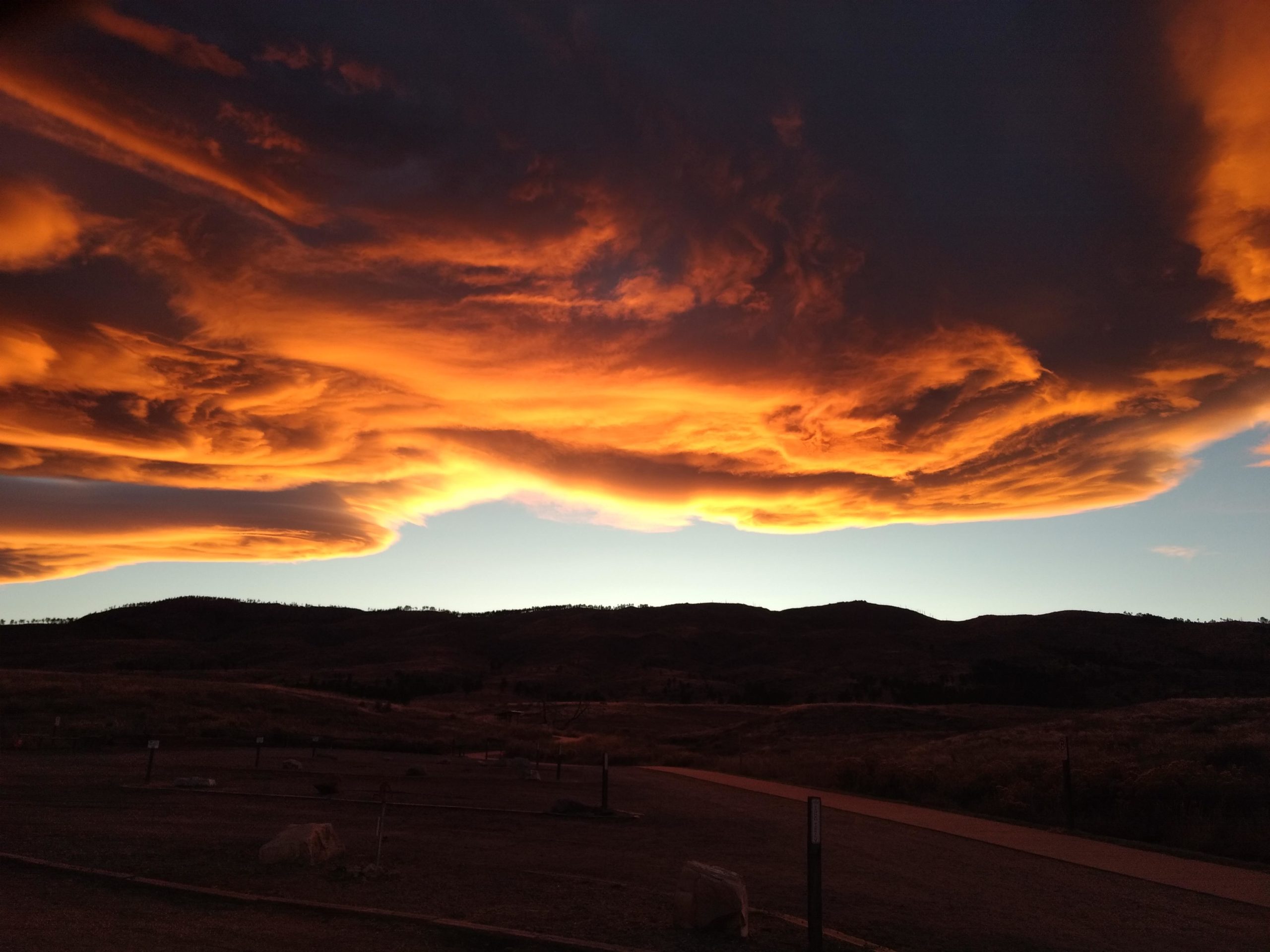A vibrant sunset sky featuring swirling orange and yellow clouds above dark silhouettes of mountains. The landscape is tranquil, with a clear outline of rolling hills and a faint blue sky at the horizon. Bobcat Ridge mountain bike trail.