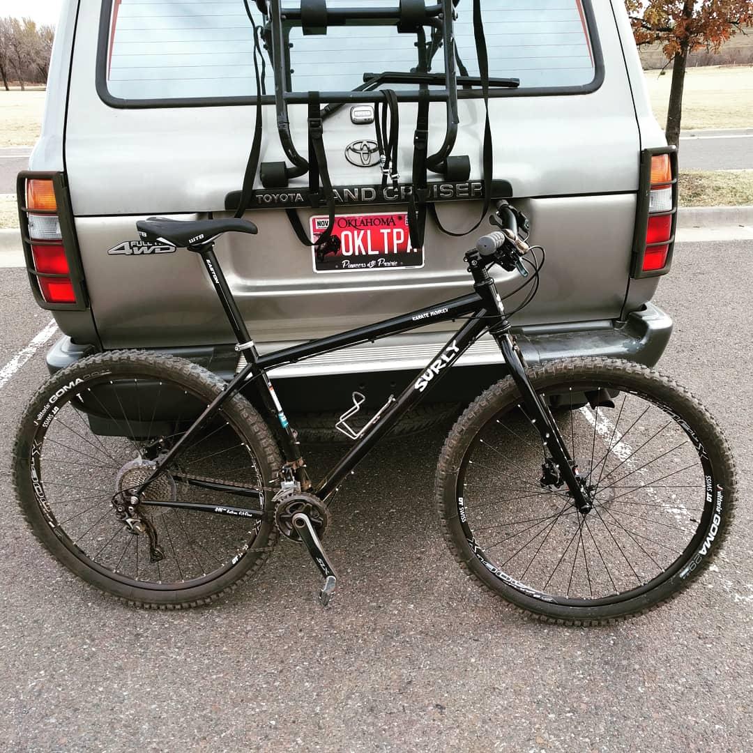 Surly Karate Monkey: A black Surly mountain bike parked next to a gray Toyota Land Cruiser. The bike is positioned at an angle, showcasing its frame and tires. The vehicle has a bike rack attached and a license plate visible with the text "OKLTP." The background features a paved area and trees in the distance.