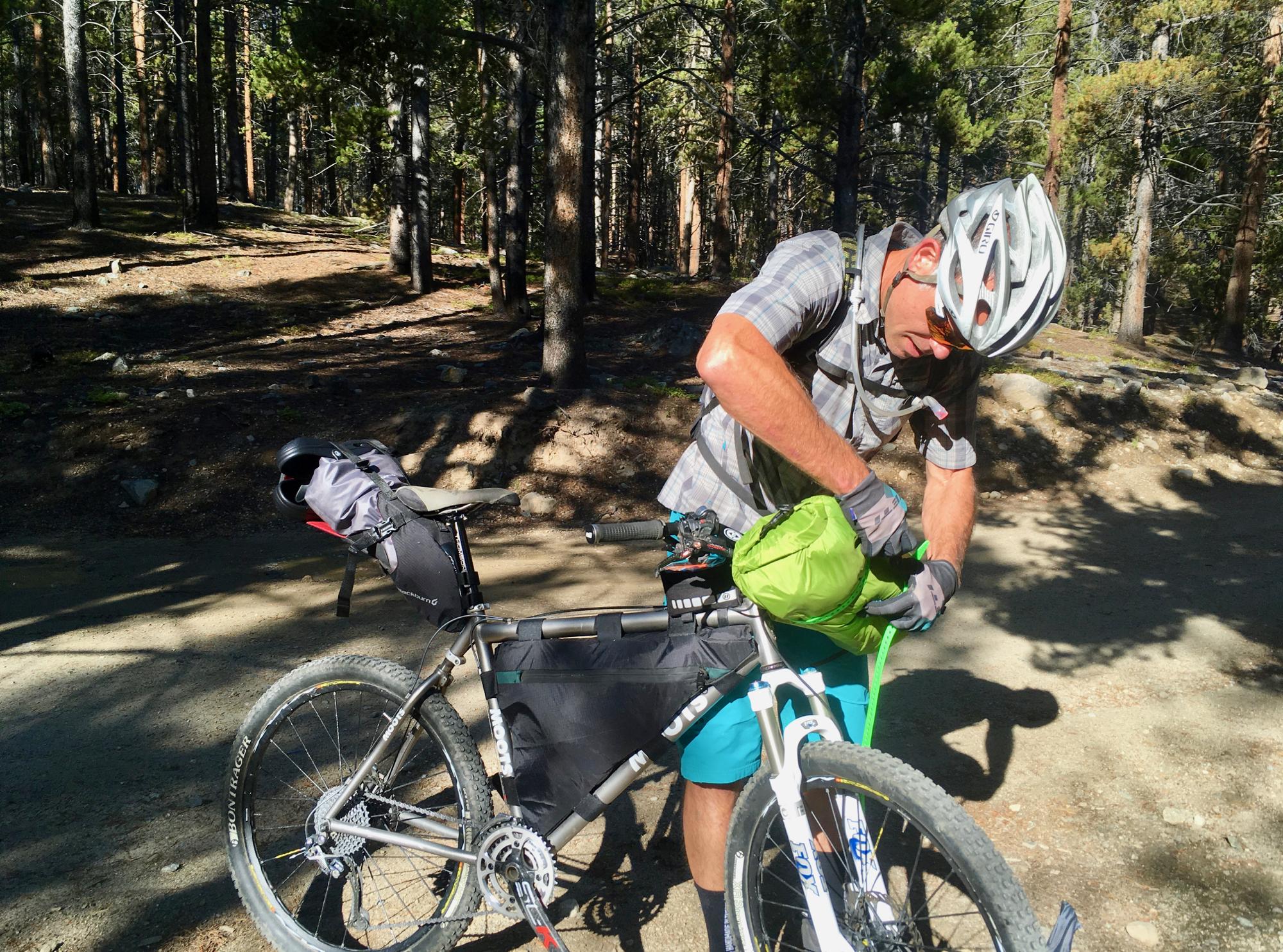A person wearing a helmet and sunglasses is adjusting a green bag on a mountain bike in a forested area. The bike has various accessories attached, and sunlight filters through the trees, creating a dappled light effect on the ground. Colorado Trail: Tennessee Pass to Holy Cross Wilderness mountain bike trail.