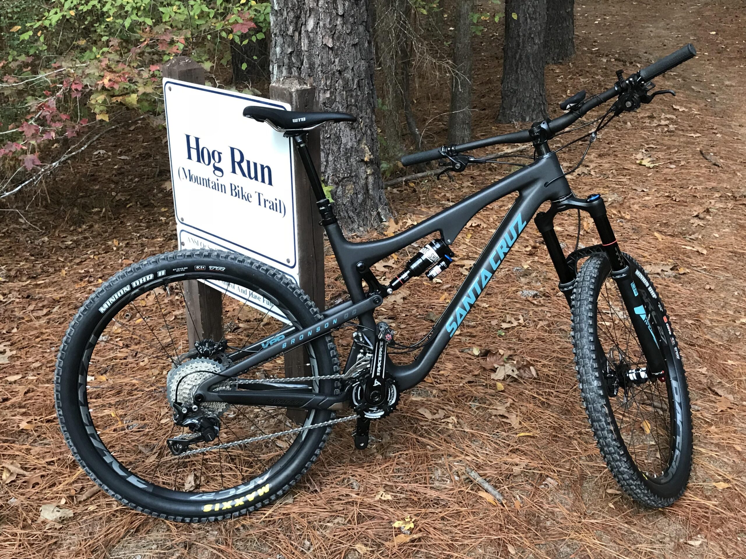Santa Cruz Bronson: A black mountain bike with blue accents leaning against a trail sign reading "Hog Run (Mountain Bike Trail)" surrounded by pine needles and trees.