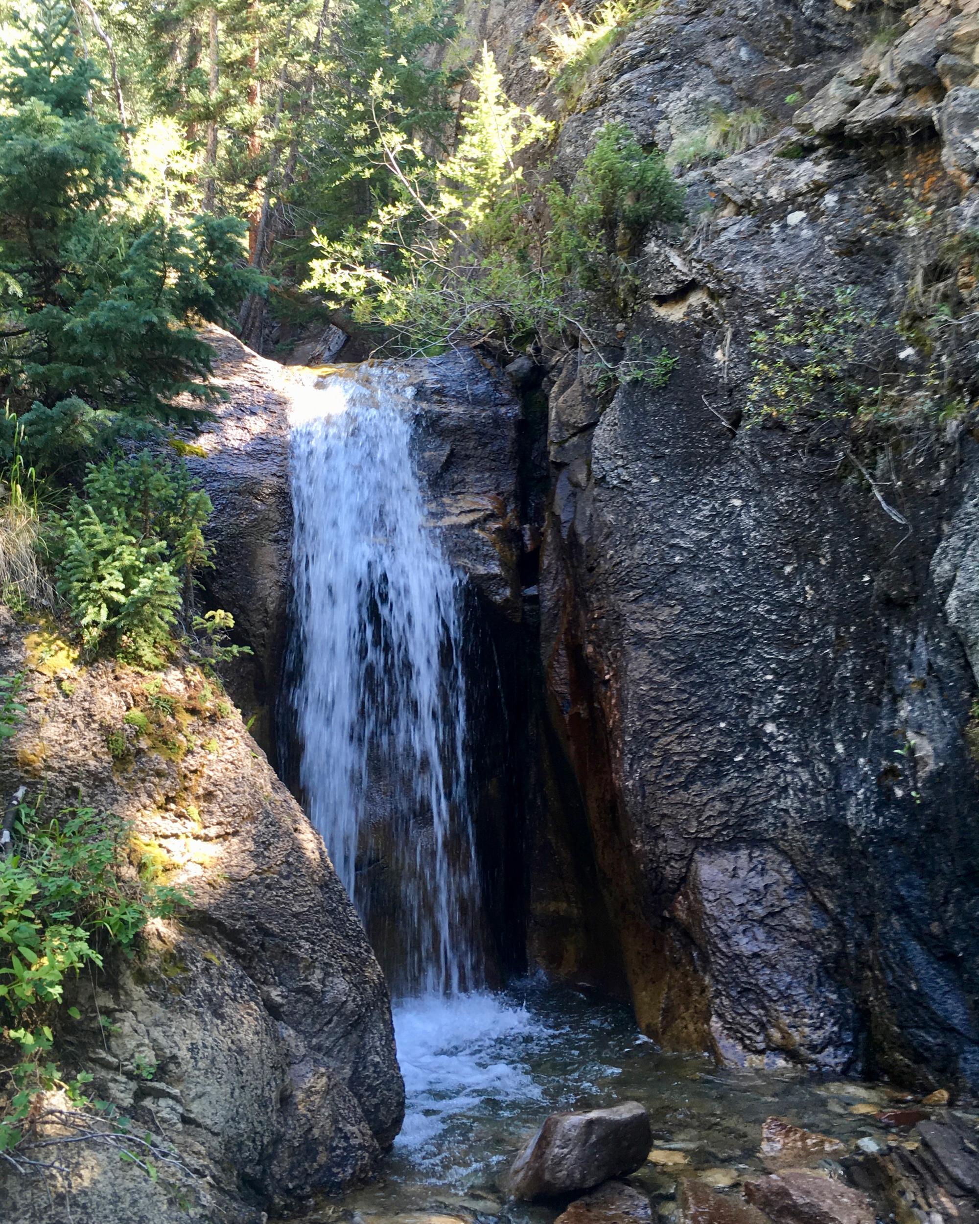 A serene waterfall cascading over rocky terrain, surrounded by lush green foliage and trees. The sunlight filters through the leaves, creating dappled light on the water and rocks. Colorado Trail: Searle Pass and Kokomo Pass (Copper Mountain to Camp Hale) mountain bike trail.