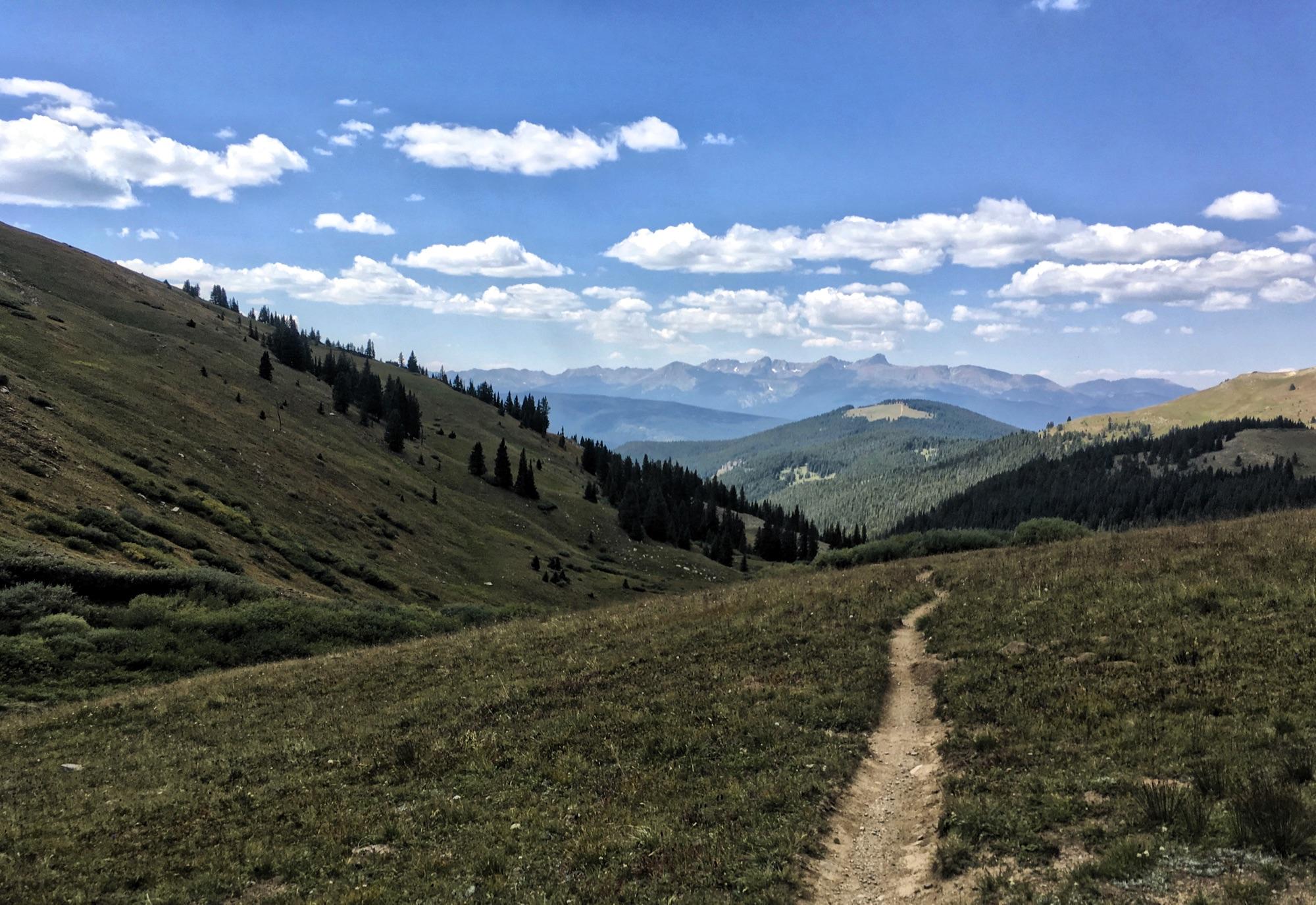A scenic view of a meandering dirt path through lush green grass and gentle hills, surrounded by tall coniferous trees under a bright blue sky with scattered clouds. Distant mountains can be seen in the background, creating a tranquil and picturesque landscape. Colorado Trail: Searle Pass and Kokomo Pass (Copper Mountain to Camp Hale) mountain bike trail.