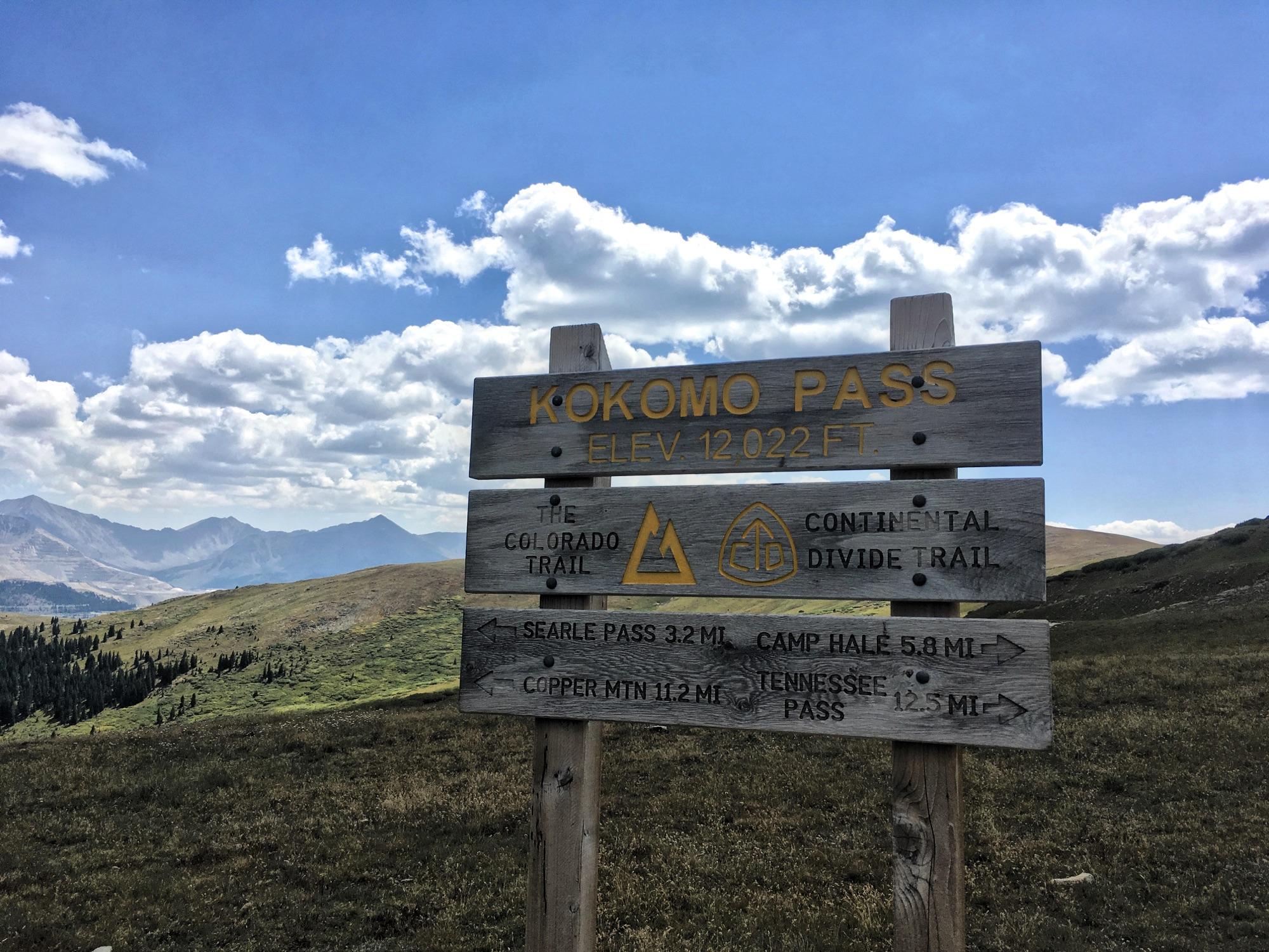Sign at Kokomo Pass, elevation 12,022 feet, displaying trail information for the Colorado Trail and Continental Divide Trail, with directional arrows indicating distances to nearby locations such as Searle Pass, Camp Hale, Copper Mountain, and Tennessee Pass, set against a backdrop of mountains and a blue sky with clouds. Colorado Trail: Searle Pass and Kokomo Pass (Copper Mountain to Camp Hale) mountain bike trail.