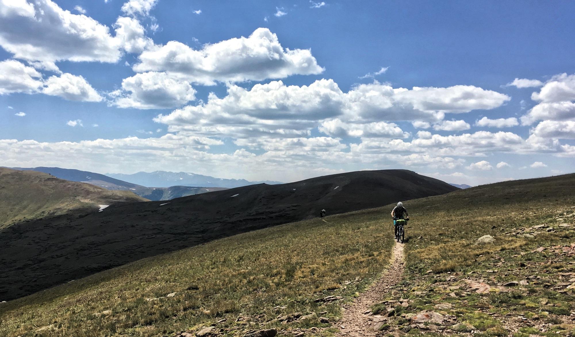 A mountain biker rides along a narrow trail through grassy terrain with rolling hills under a blue sky dotted with fluffy clouds. Mountains are visible in the background, creating a picturesque outdoor scene. Colorado Trail: Searle Pass and Kokomo Pass (Copper Mountain to Camp Hale) mountain bike trail.