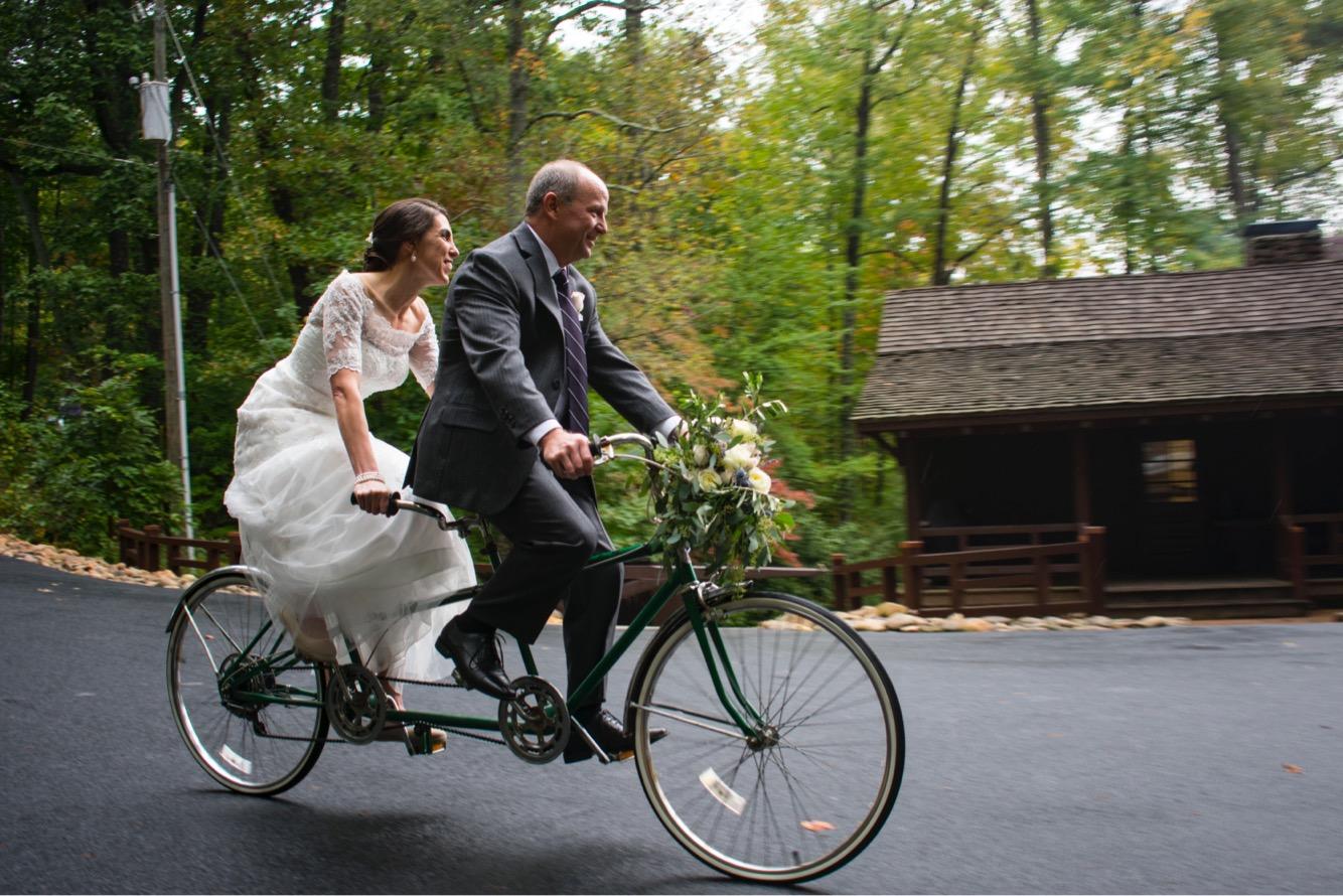 A happy couple riding a tandem bicycle, with the bride in a white wedding dress and the groom in a gray suit. They are surrounded by lush greenery, and the scene captures a joyful and playful moment during their wedding day. The bicycle is adorned with floral decorations, and a rustic cabin can be seen in the background. Paris Mountain State Park mountain bike trail.
