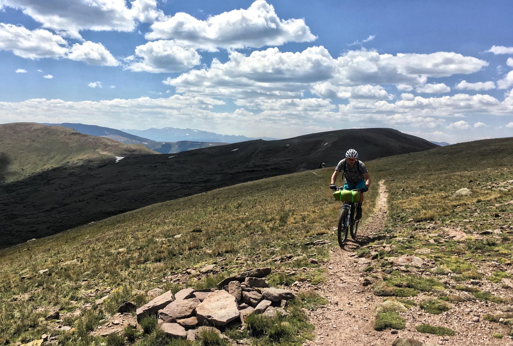 A cyclist riding a mountain bike along a narrow dirt trail in a mountainous landscape, surrounded by green hills and a blue sky with white clouds. In the foreground, there are rocks along the trail, and additional hills and distant mountains are visible in the background. Colorado Trail: Searle Pass and Kokomo Pass (Copper Mountain to Camp Hale) mountain bike trail.