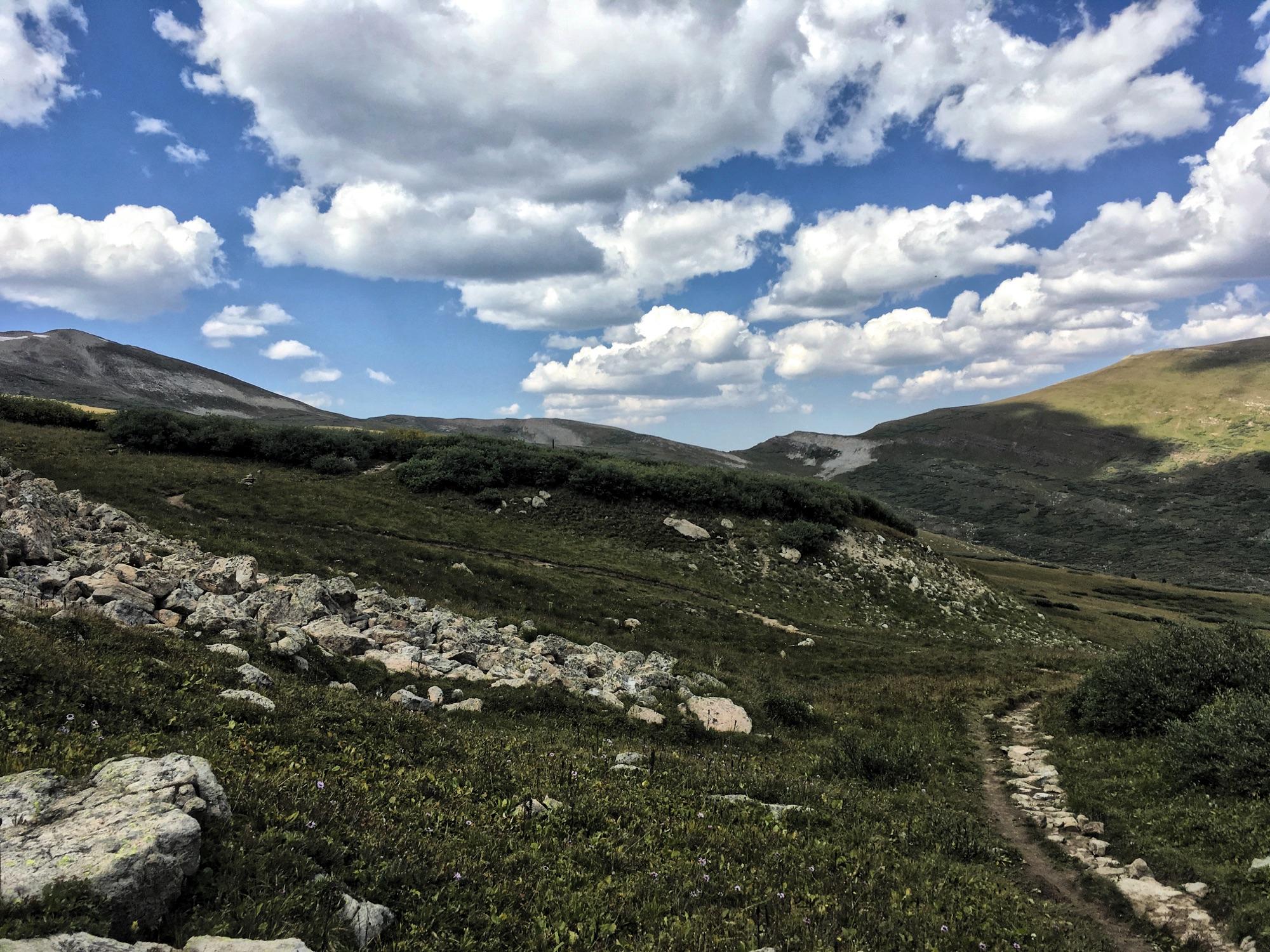A scenic landscape featuring rolling hills and rocky terrain under a partly cloudy sky. A winding dirt path leads through lush green grass and scattered rocks, with distant mountains visible in the background. Colorado Trail: Searle Pass and Kokomo Pass (Copper Mountain to Camp Hale) mountain bike trail.