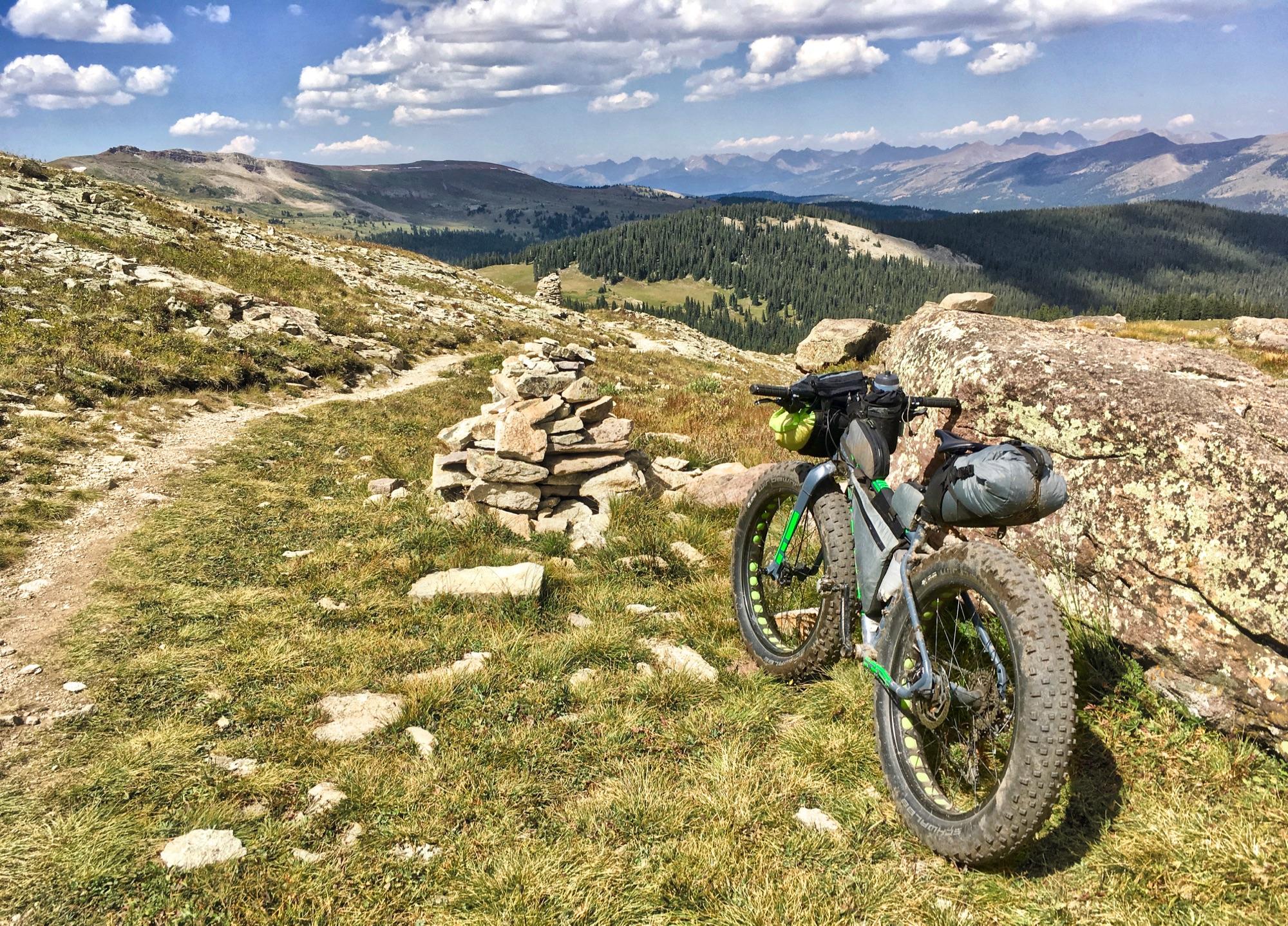 A fat tire bicycle parked beside a rock stack on a mountain trail, with a panoramic view of rolling hills and distant mountains under a partly cloudy sky. The grassy terrain is dotted with scattered rocks, and a winding path leads into the landscape. Colorado Trail: Searle Pass and Kokomo Pass (Copper Mountain to Camp Hale) mountain bike trail.