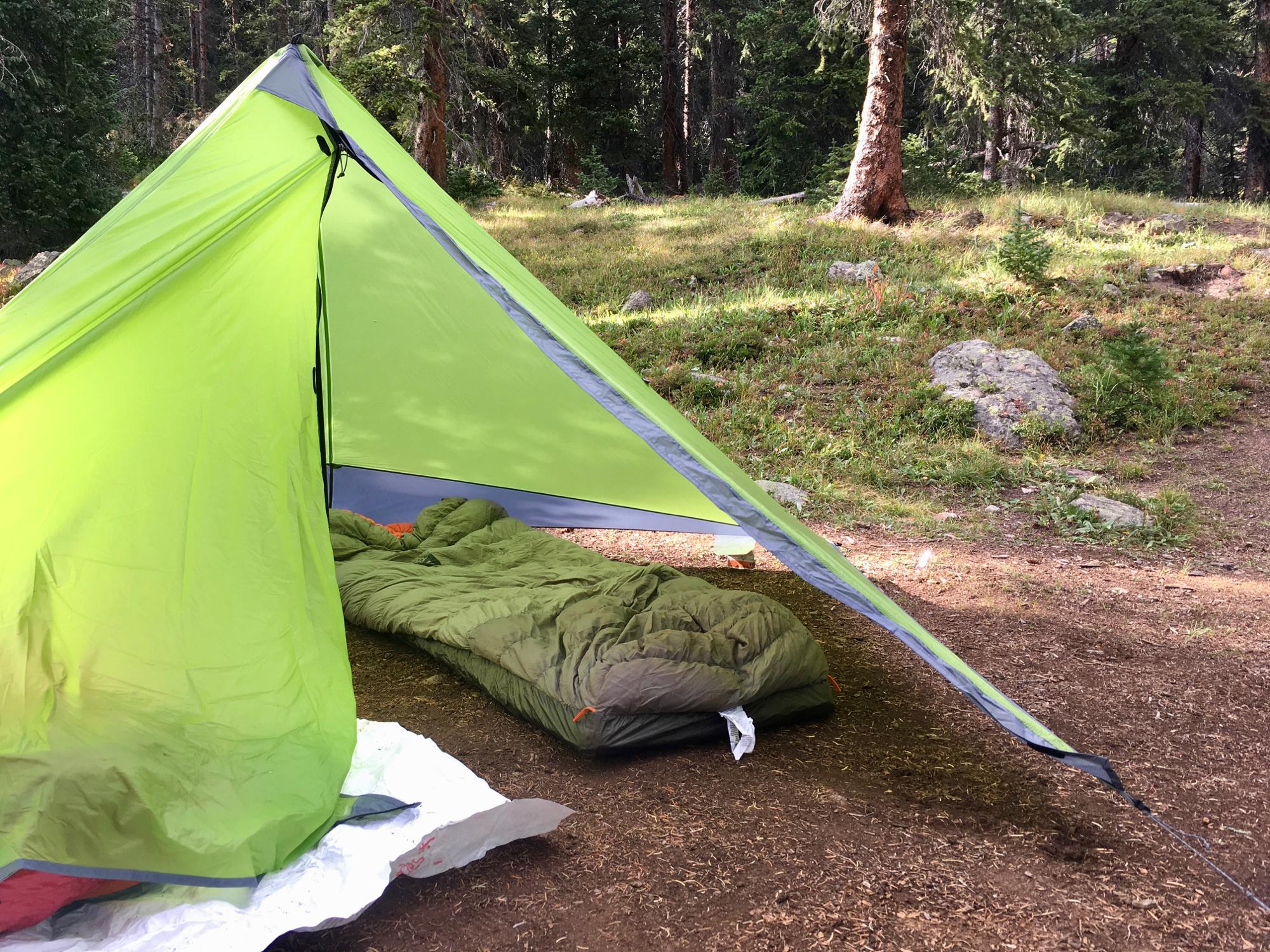 A green camping tent set up in a wooded area, with a sleeping bag partially visible inside. The ground is covered in brown earth and patches of grass, with trees in the background. Colorado Trail: Searle Pass and Kokomo Pass (Copper Mountain to Camp Hale) mountain bike trail.