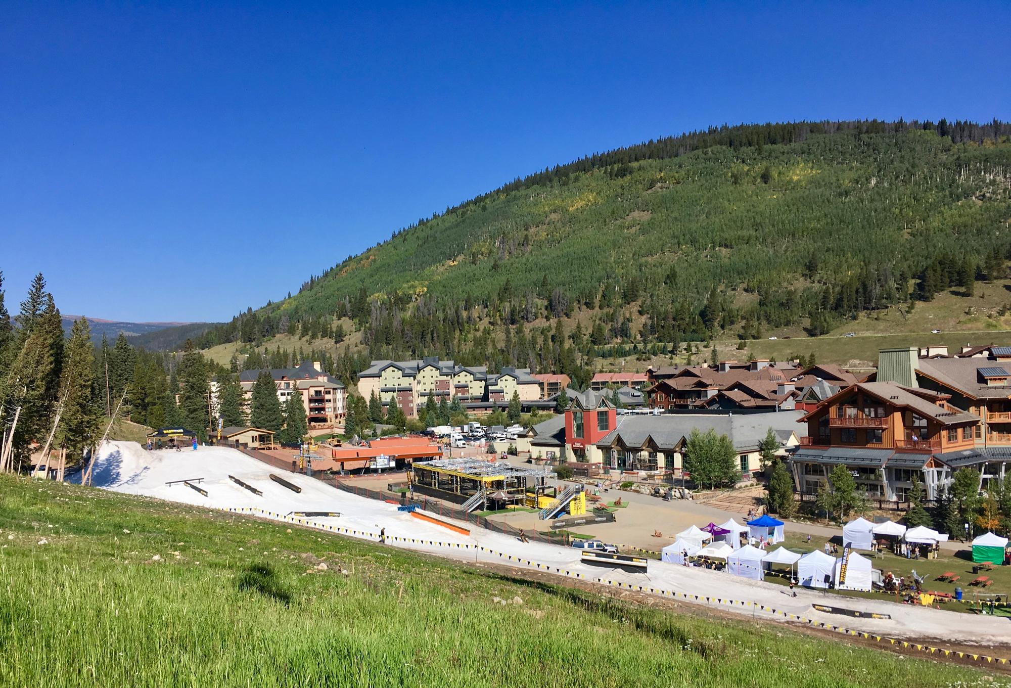 A scenic view of a mountainous area featuring lush green hills and a clear blue sky. In the foreground, a ski slope with various terrain features is visible, alongside a grassy area. Below, a collection of buildings includes residential lodgings and commercial spaces, with several tents set up for an event. The backdrop showcases more green mountains, indicating a vibrant, outdoor recreational environment. Colorado Trail: Searle Pass and Kokomo Pass (Copper Mountain to Camp Hale) mountain bike trail.