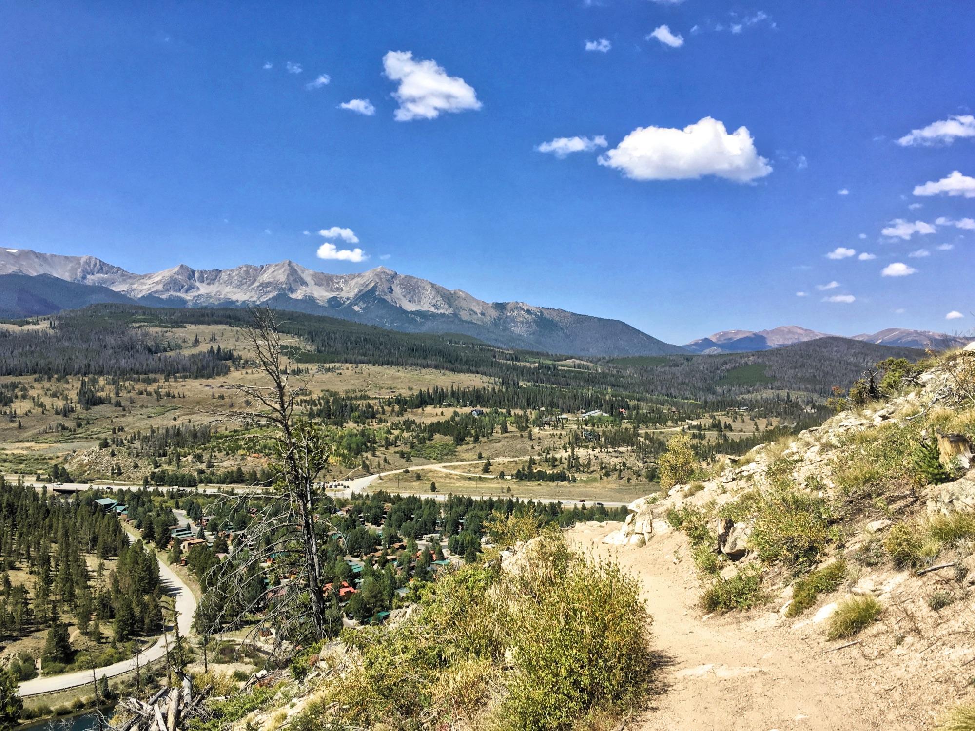 A panoramic view of a mountainous landscape under a clear blue sky, featuring rugged peaks in the background, rolling hills, and a mix of evergreen trees and open land. In the foreground, a dirt path leads through light vegetation, while a small town is visible nestled in the valley below. Fluffy white clouds dot the sky, adding to the serene natural scene. Colorado Trail: Kenosha Pass To Breckenridge mountain bike trail.