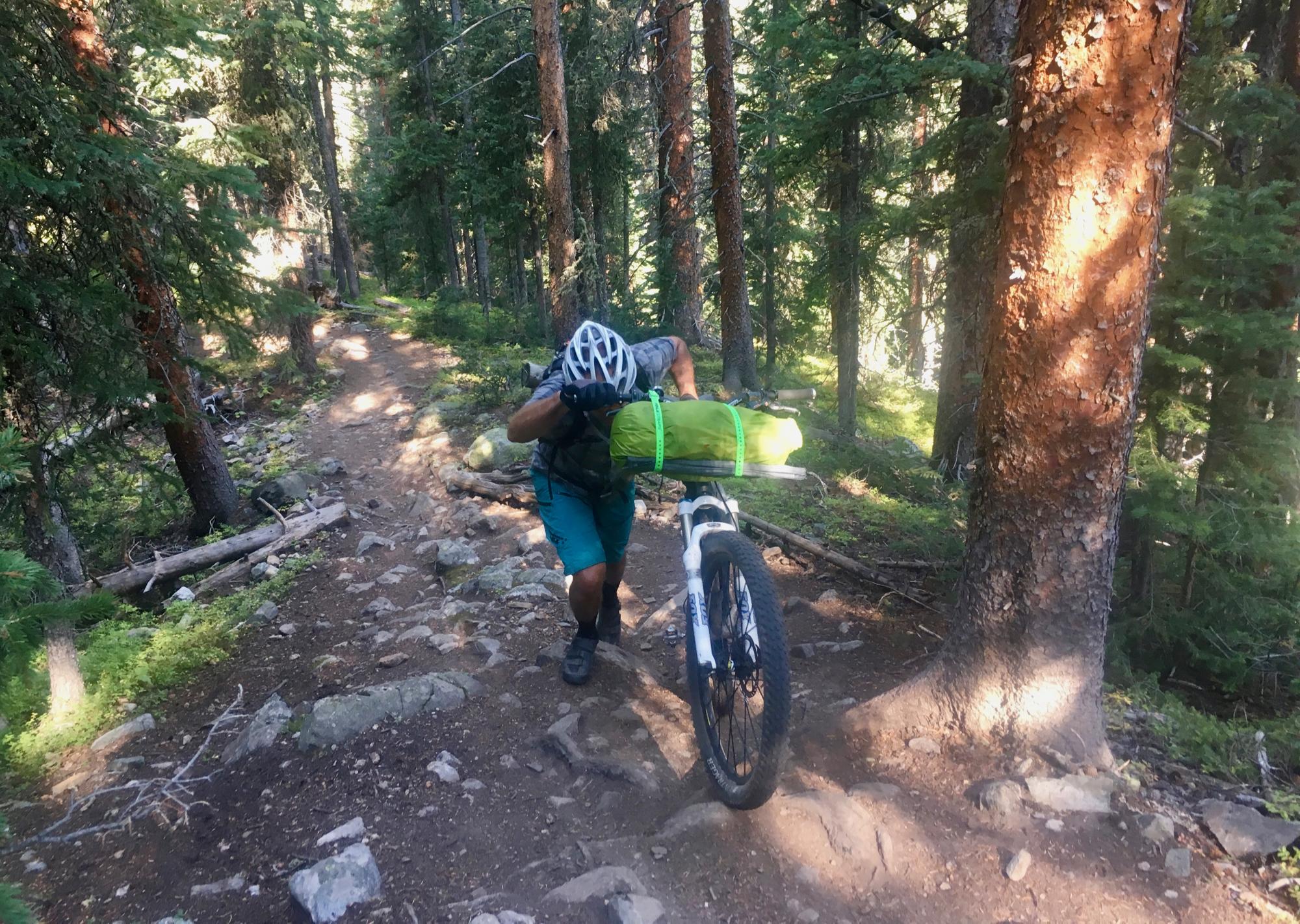A cyclist pushing a mountain bike along a rocky trail in a dense forest, with tall trees and sunlight filtering through the leaves. The cyclist is wearing a helmet and has a green bag strapped to the bike. Colorado Trail: Kenosha Pass To Breckenridge mountain bike trail.
