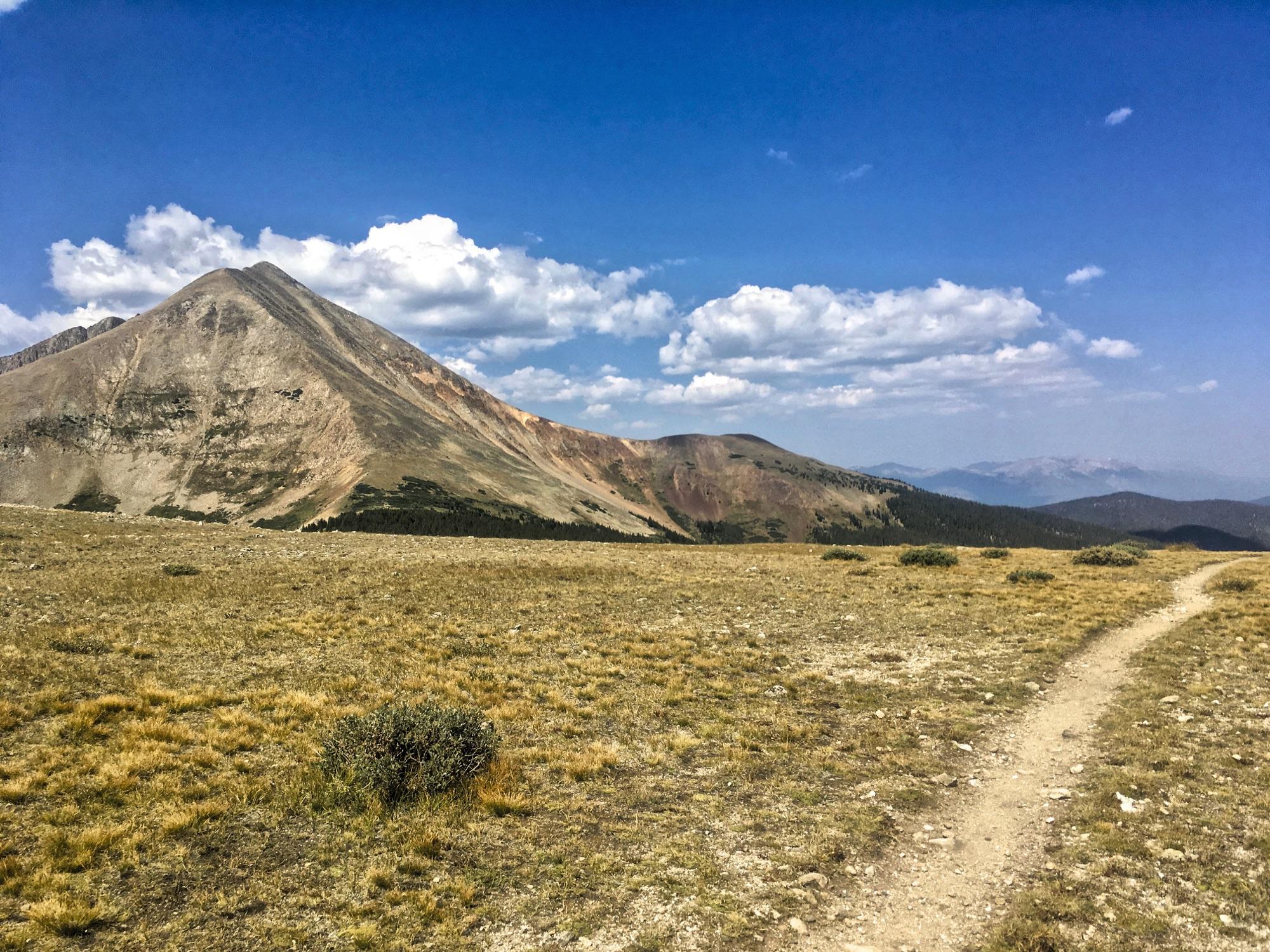 A scenic landscape featuring a rugged mountain peak under a bright blue sky with scattered clouds. In the foreground, a dirt trail meanders through a grassy field, leading towards the mountains in the background. The terrain is marked by rocky outcrops and patches of vegetation, creating a serene and natural setting. Colorado Trail: Kenosha Pass To Breckenridge mountain bike trail.