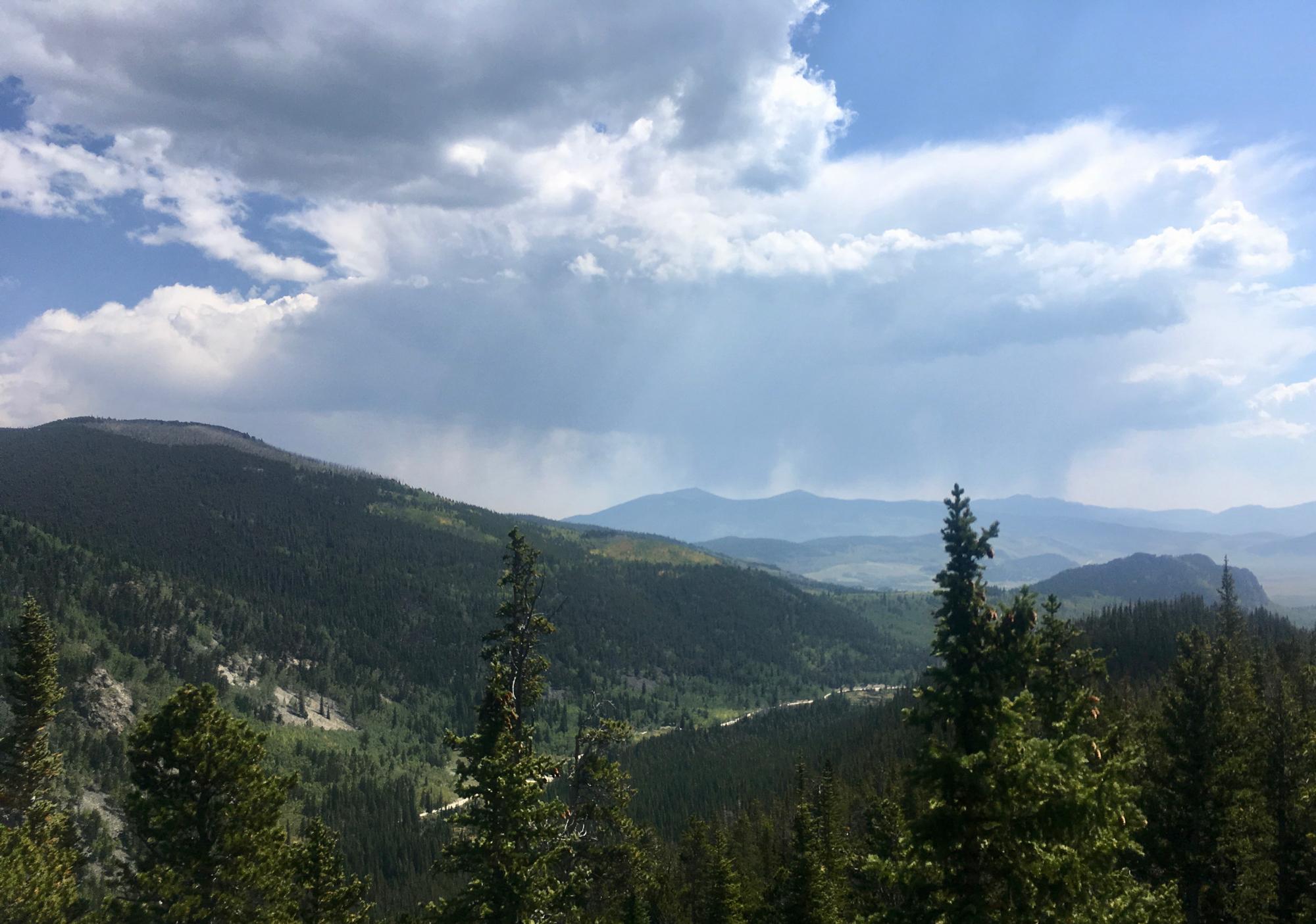 A scenic landscape featuring rolling green hills and mountains under a partly cloudy sky, with patches of blue and wisps of clouds. Pine trees are visible in the foreground, while the distant mountains fade into a hazy background, suggesting a vast natural setting. Colorado Trail: Kenosha Pass To Breckenridge mountain bike trail.