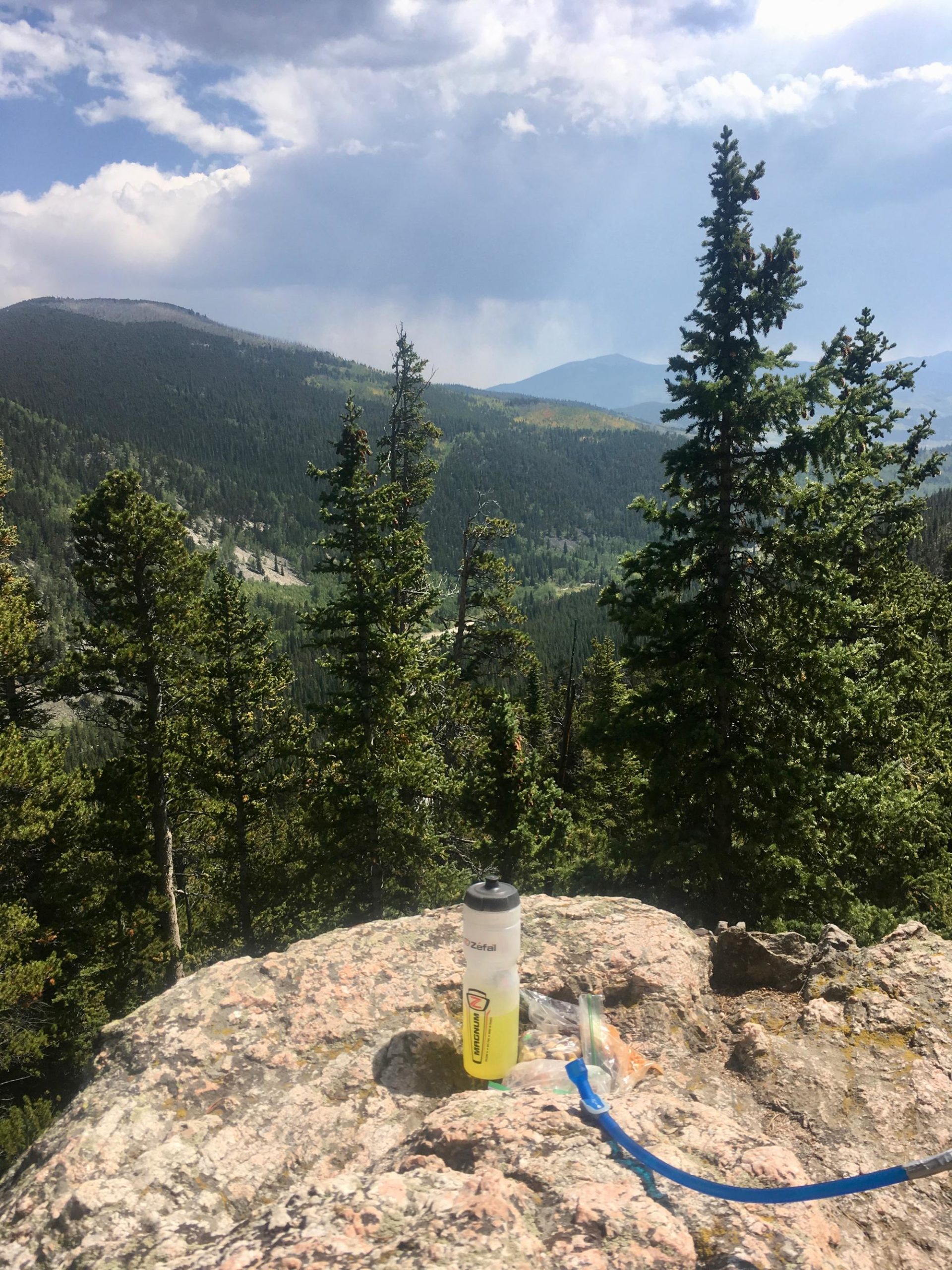 A scenic view of a mountainous landscape with lush green forests and distant peaks under a partly cloudy sky. In the foreground, a clear rock surface features a yellow water bottle and a hydration tube, suggesting a location for rest and refreshment during a hike. Colorado Trail: Kenosha Pass To Breckenridge mountain bike trail.