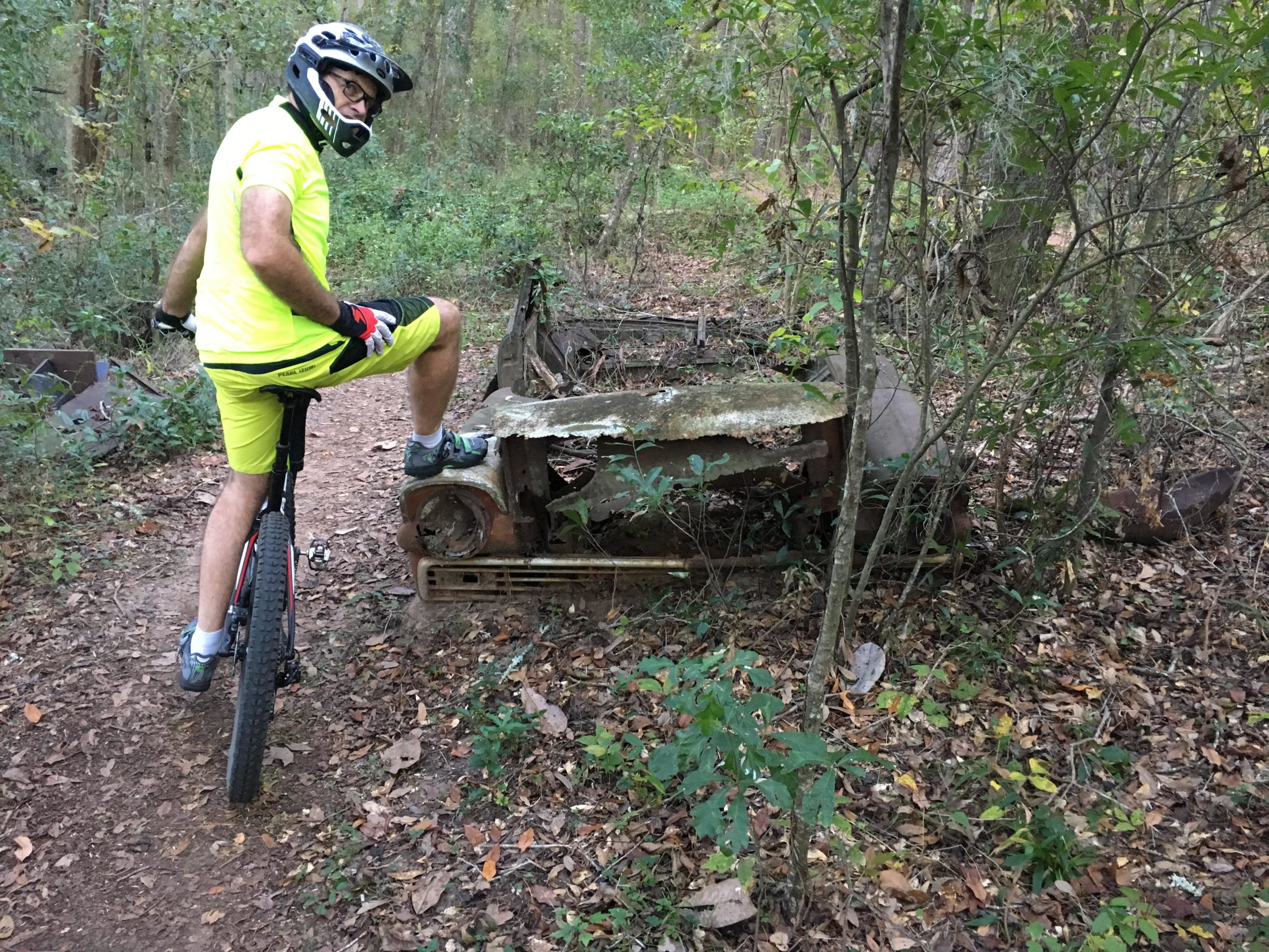 A person wearing a bright yellow cycling outfit and helmet is balancing on a mountain bike near an abandoned, rusty vehicle in a wooded area. The trail is surrounded by greenery and fallen leaves. Cadillac mountain bike trail.
