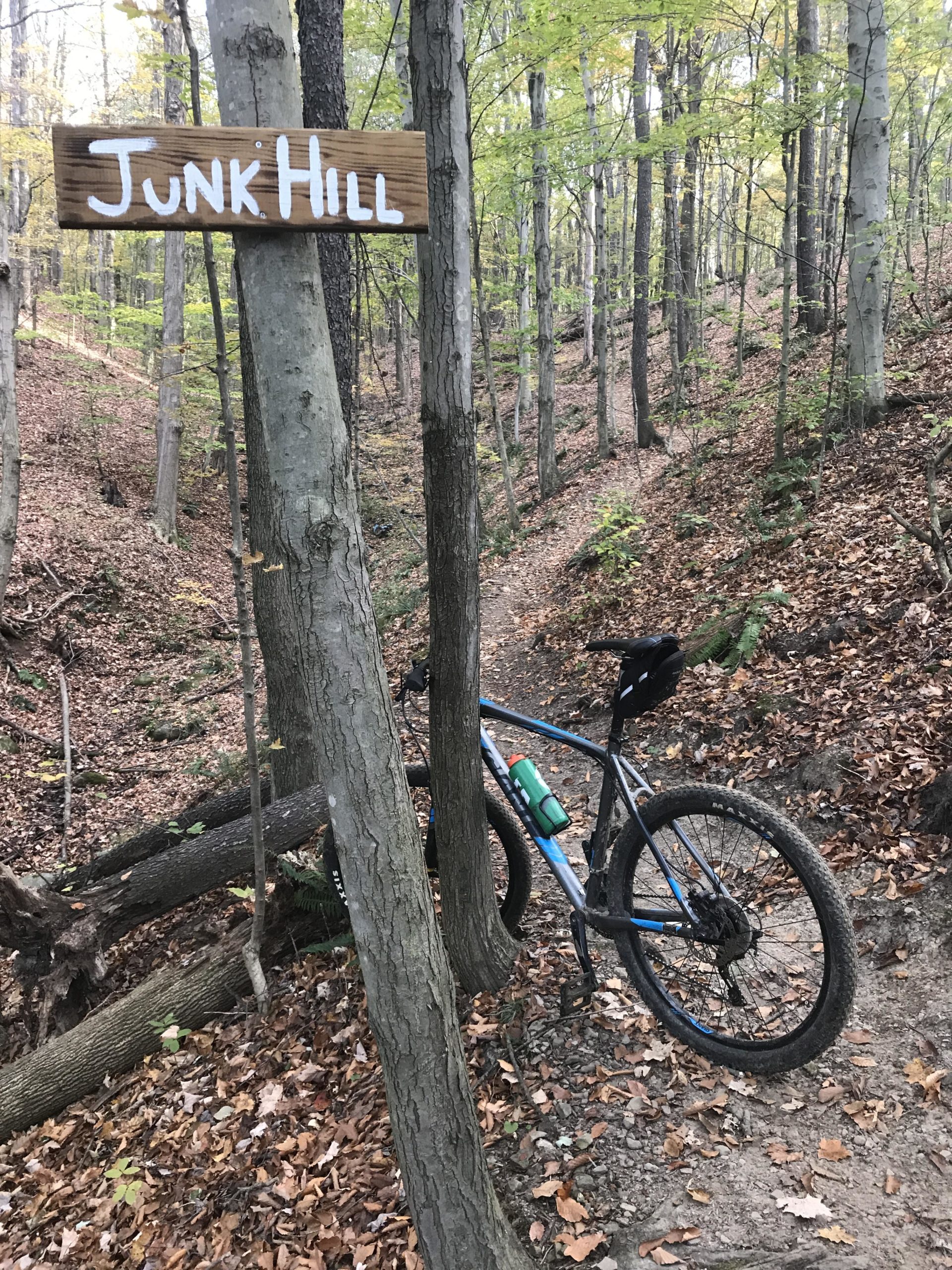 A mountain bike is leaning against a tree next to a wooden sign that reads "Junk Hill." The scene is in a forested area with trees showing hints of fall foliage and a trail visible through the fallen leaves on the ground. Vultures Knob mountain bike trail.