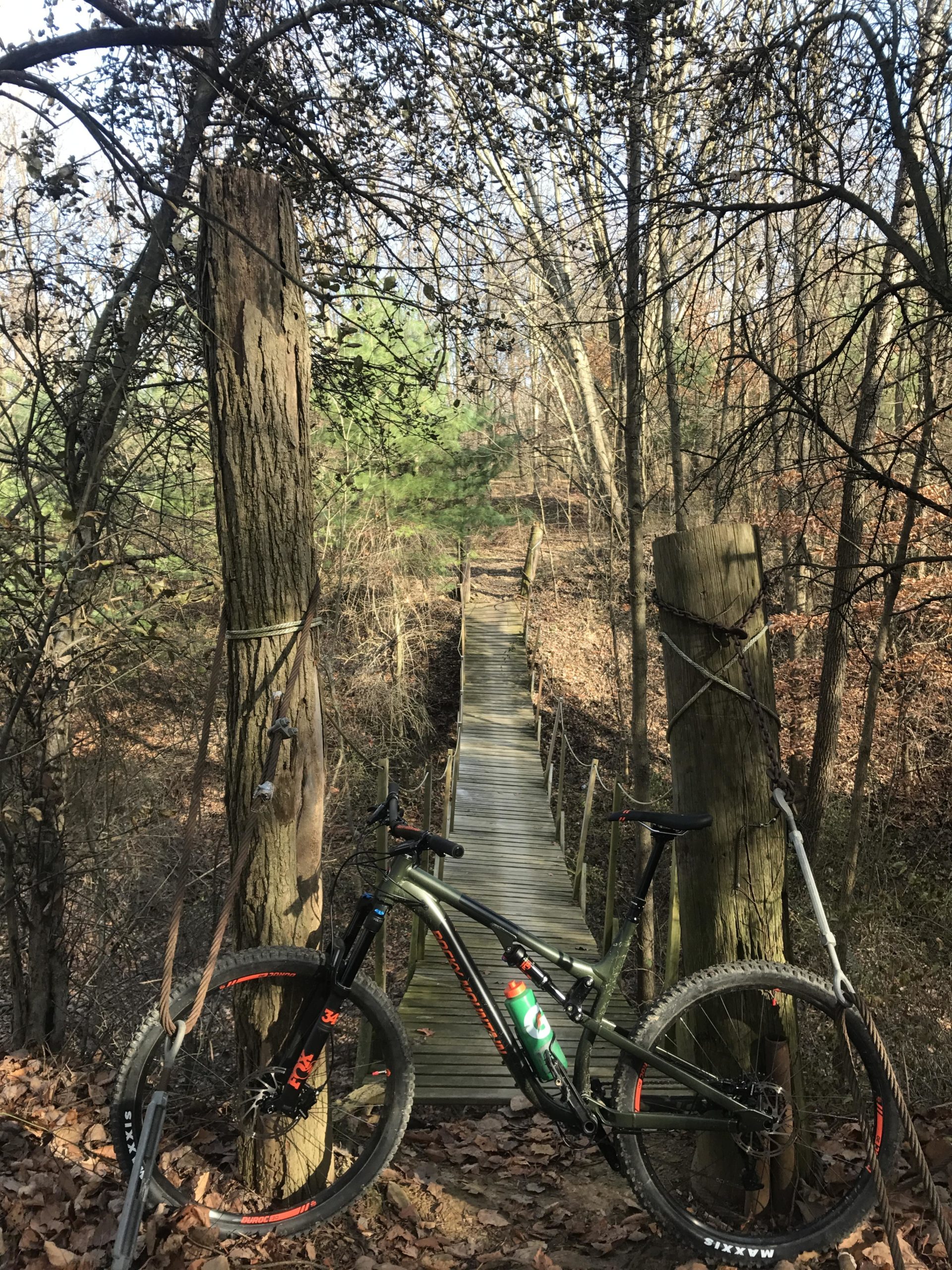 A mountain bike rests on the ground beside two wooden posts at the edge of a narrow bridge. The bridge, made of wooden planks, leads across a small ravine surrounded by bare trees and sparse underbrush. Sunlight filters through the branches, illuminating the scene in a calm, natural setting. Vultures Knob mountain bike trail.