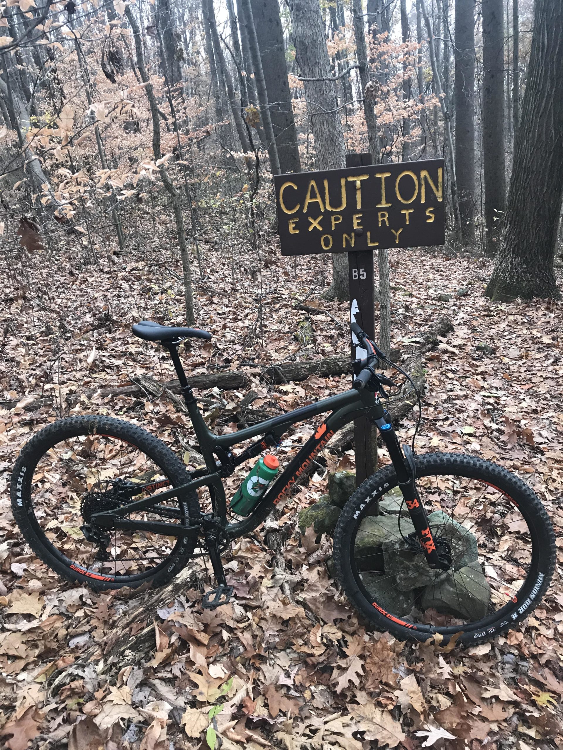 A mountain bike resting on the ground near a "Caution: Experts Only" sign in a wooded area. The scene features autumn leaves scattered on the ground and trees in the background, indicating a natural trail environment. A water bottle is attached to the bike, suggesting preparation for a ride. West Branch mountain bike trail.