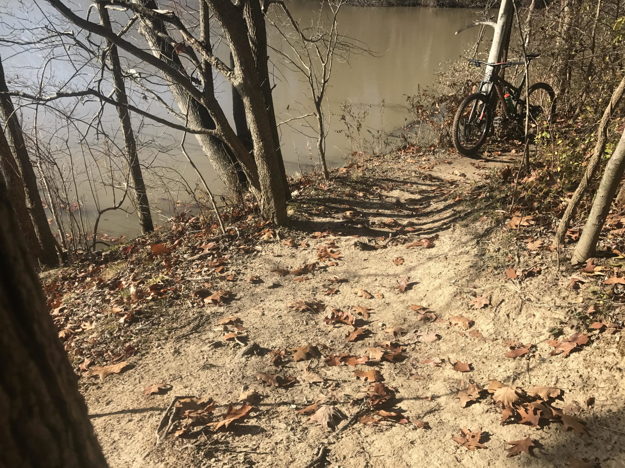 A narrow dirt path lined with fallen leaves leads towards a calm river. In the background, trees frame the water's edge, and a bicycle is parked nearby, partially obscured by the foliage. The scene captures a tranquil outdoor setting, showcasing autumn colors and a hint of nature's serenity. Findley State Park mountain bike trail.
