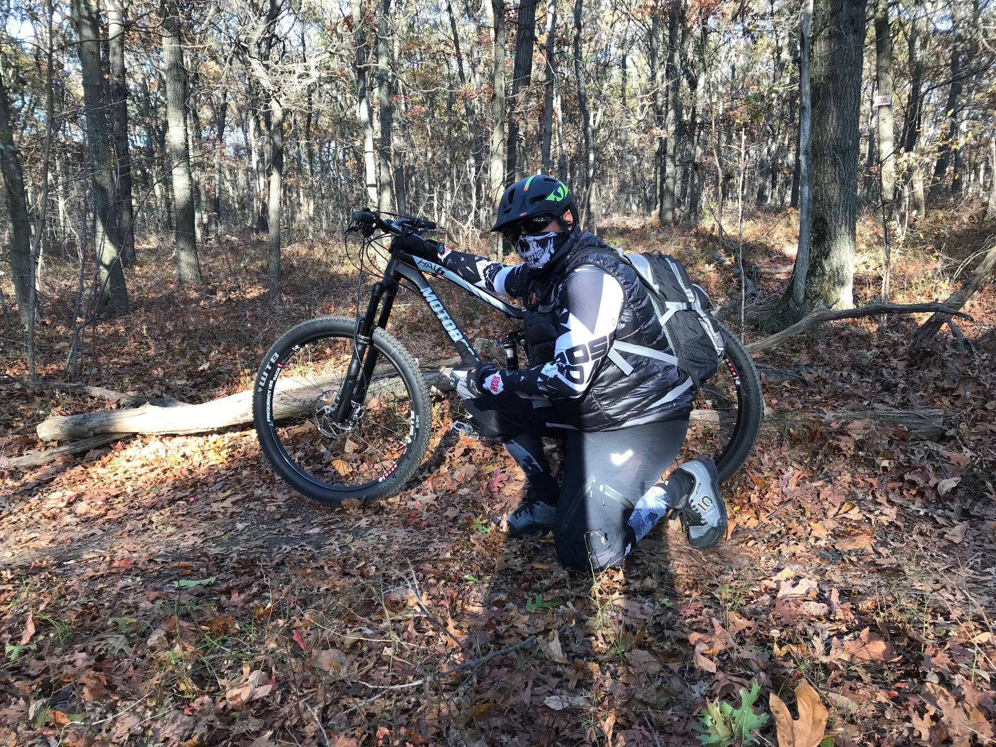 A person wearing a helmet and a face covering crouches beside a mountain bike in a forested area with autumn leaves on the ground. The cyclist is dressed in sports gear, including a vest and padded shorts, and appears to be examining the bike. Sunlight filters through the trees, creating a natural and serene outdoor setting. Trail View State Park mountain bike trail.
