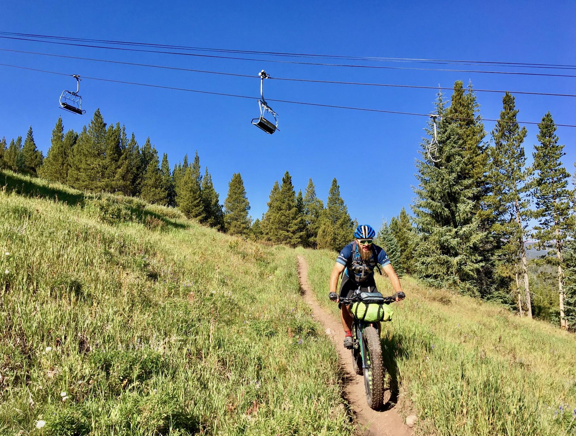 A mountain biker rides along a grassy trail surrounded by trees, with ski lift chairs visible overhead against a blue sky. The cyclist is wearing a helmet and riding a fat bike equipped with a large handlebar bag. Colorado Trail: Searle Pass and Kokomo Pass (Copper Mountain to Camp Hale) mountain bike trail.