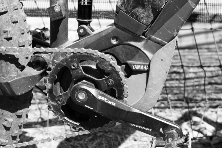 Close-up of a bicycle's crankset, chain, and motor, showcasing a dirty chain and components in black and white. The image highlights the Yamaha branding on the motor and the intricate details of the crank and gears.