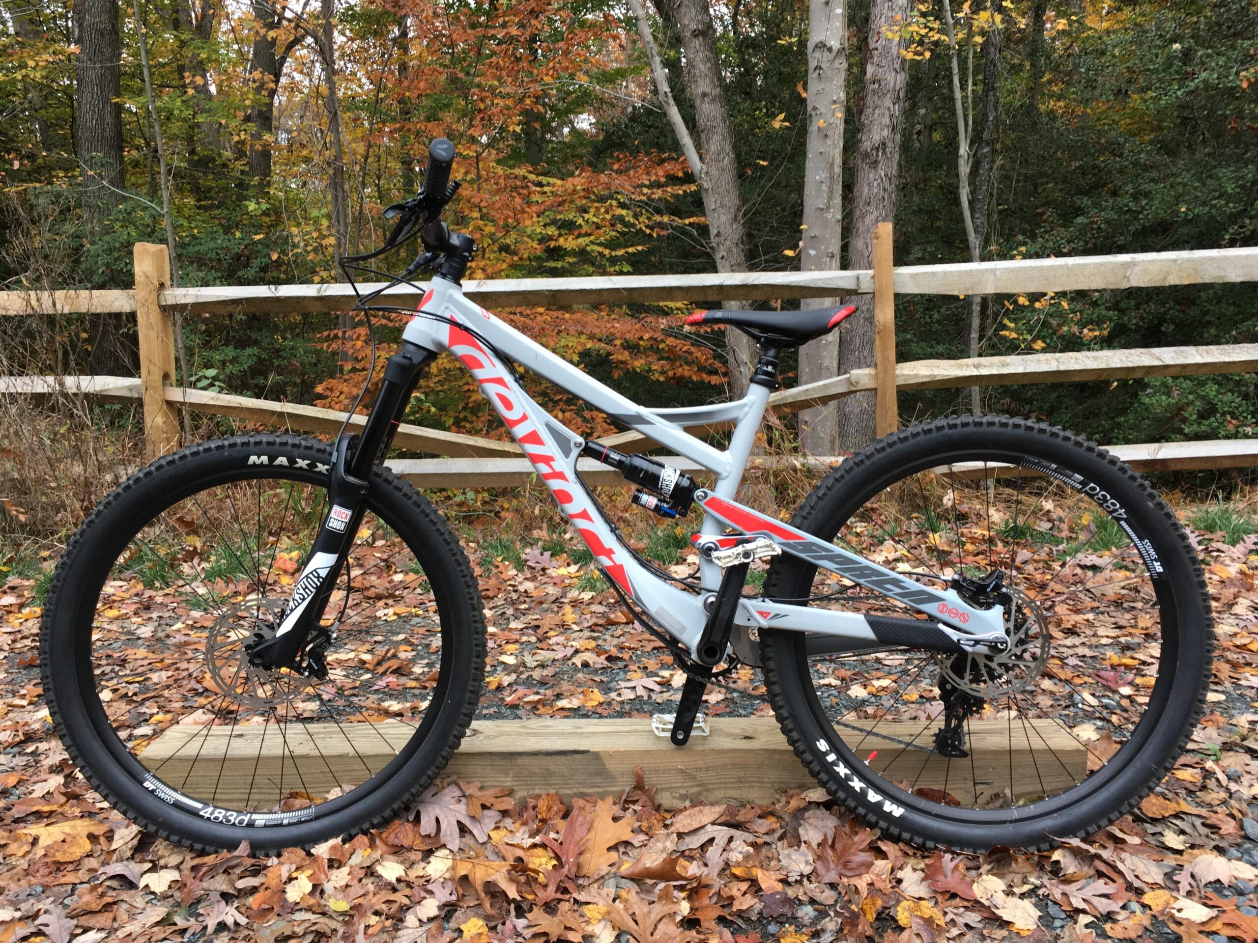 Devinci Spartan: An off-road mountain bike positioned on a wooden platform, surrounded by colorful autumn leaves and trees in the background. The bike features a gray frame with red accents and is equipped with thick tires suitable for rough terrain. A wooden fence runs parallel to the bike, enhancing the natural setting.