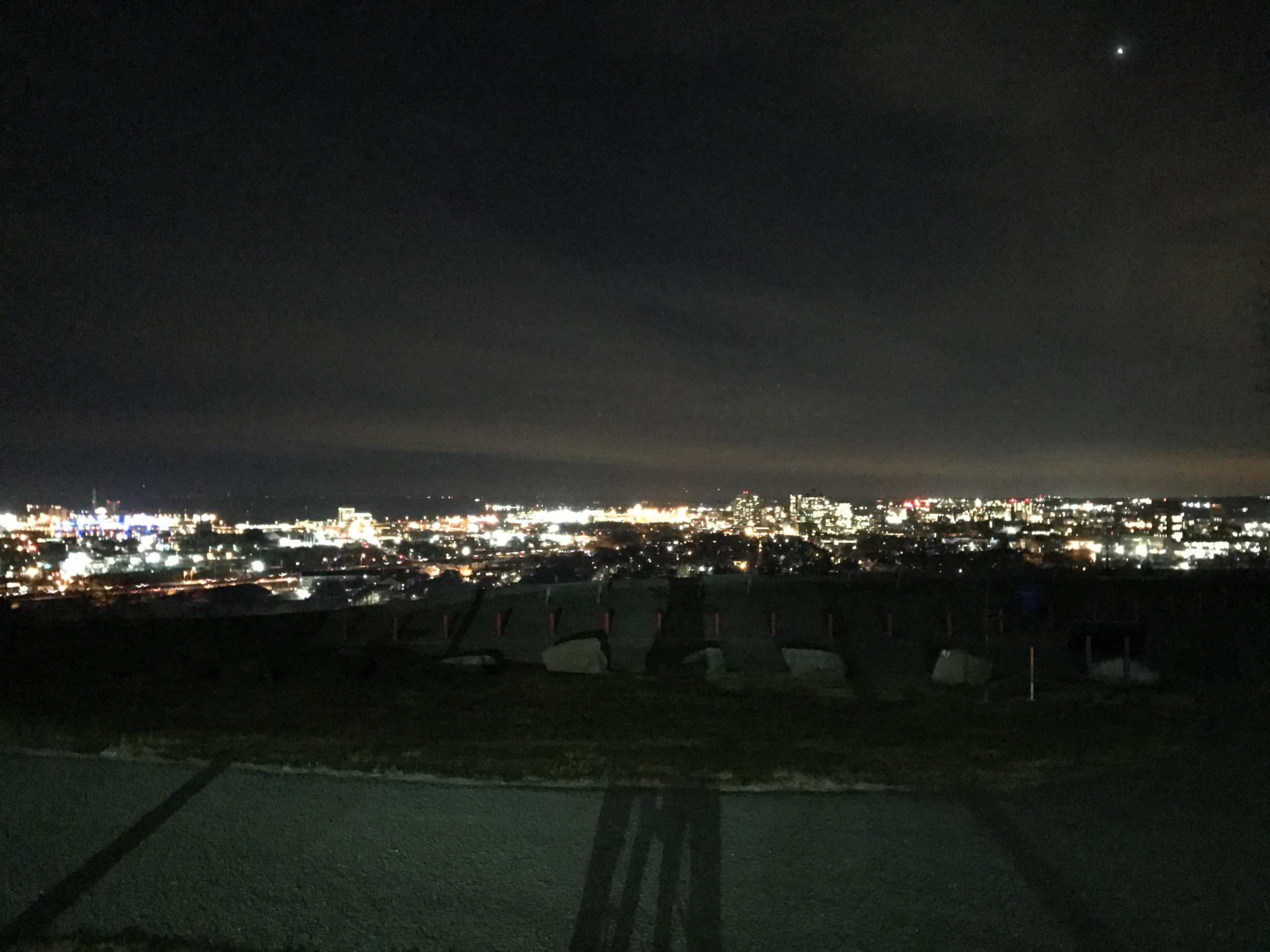 A nighttime view of a city skyline illuminated by numerous lights, with a dark sky above and a few scattered clouds. The foreground features a low hill or park area, along with shadows cast by objects or people. The distant buildings have colorful lights, and a bright star is visible in the upper right corner of the sky. East Rock Park mountain bike trail.