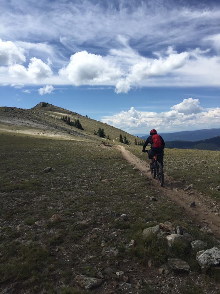 A mountain biker rides along a rugged dirt trail through a grassy landscape, with a backdrop of rolling hills and a blue sky filled with fluffy white clouds. Monarch Crest Trail mountain bike trail.