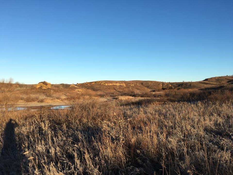 A scenic landscape featuring rolling hills under a clear blue sky, with dry grass and shrubs in the foreground and a small body of water visible in the lower left corner. The vegetation appears frost-kissed, indicating a chilly atmosphere. Kanopolis Lake mountain bike trail.