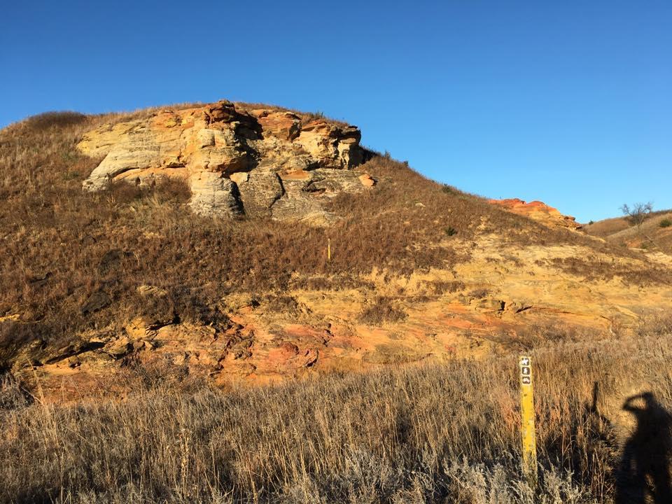A scenic view of a rocky hillside under a clear blue sky, showcasing layers of colored rock formations. The foreground features dry grass, while a yellow post with a number stands on the right side. The image captures the natural beauty of the landscape. Kanopolis Lake mountain bike trail.