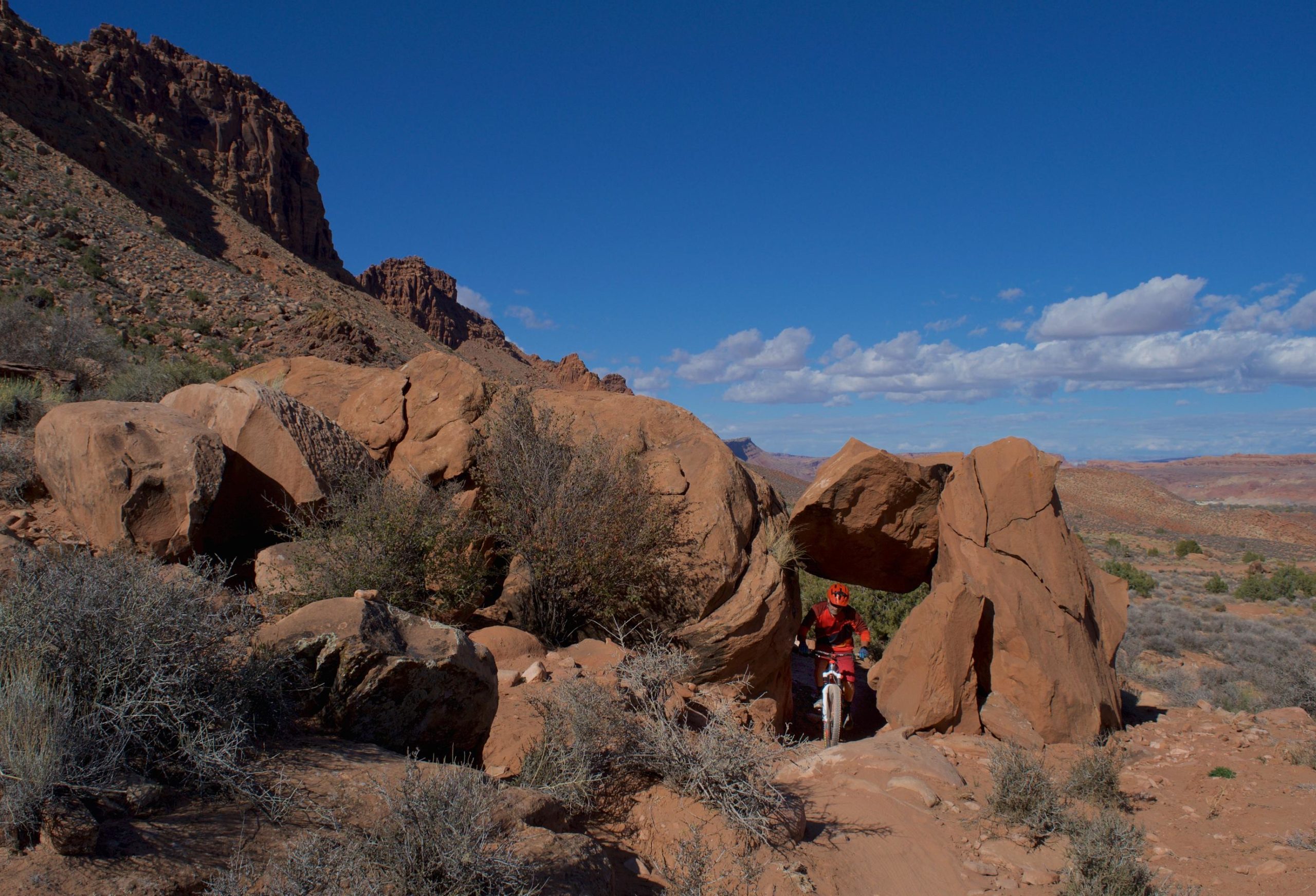 A mountain biker riding through a natural rock arch in a rugged desert landscape, surrounded by red rock formations, sparse vegetation, and a clear blue sky with a few clouds. Pipe Dream mountain bike trail.