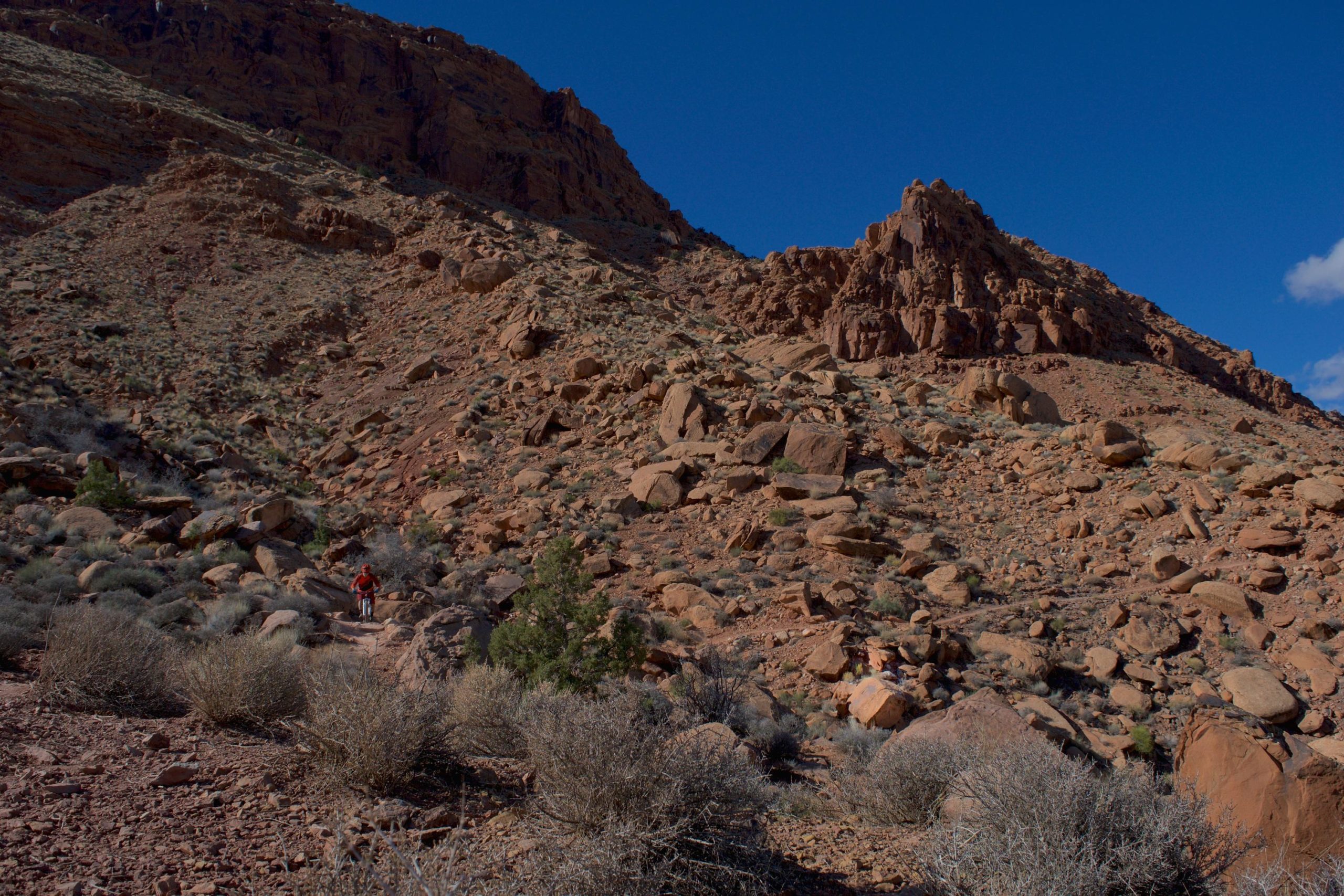 A hiker in red traverses a rocky landscape with steep hills and sparse vegetation under a clear blue sky. The terrain is comprised of large boulders and dry bushes, showcasing a rugged desert environment. Pipe Dream mountain bike trail.