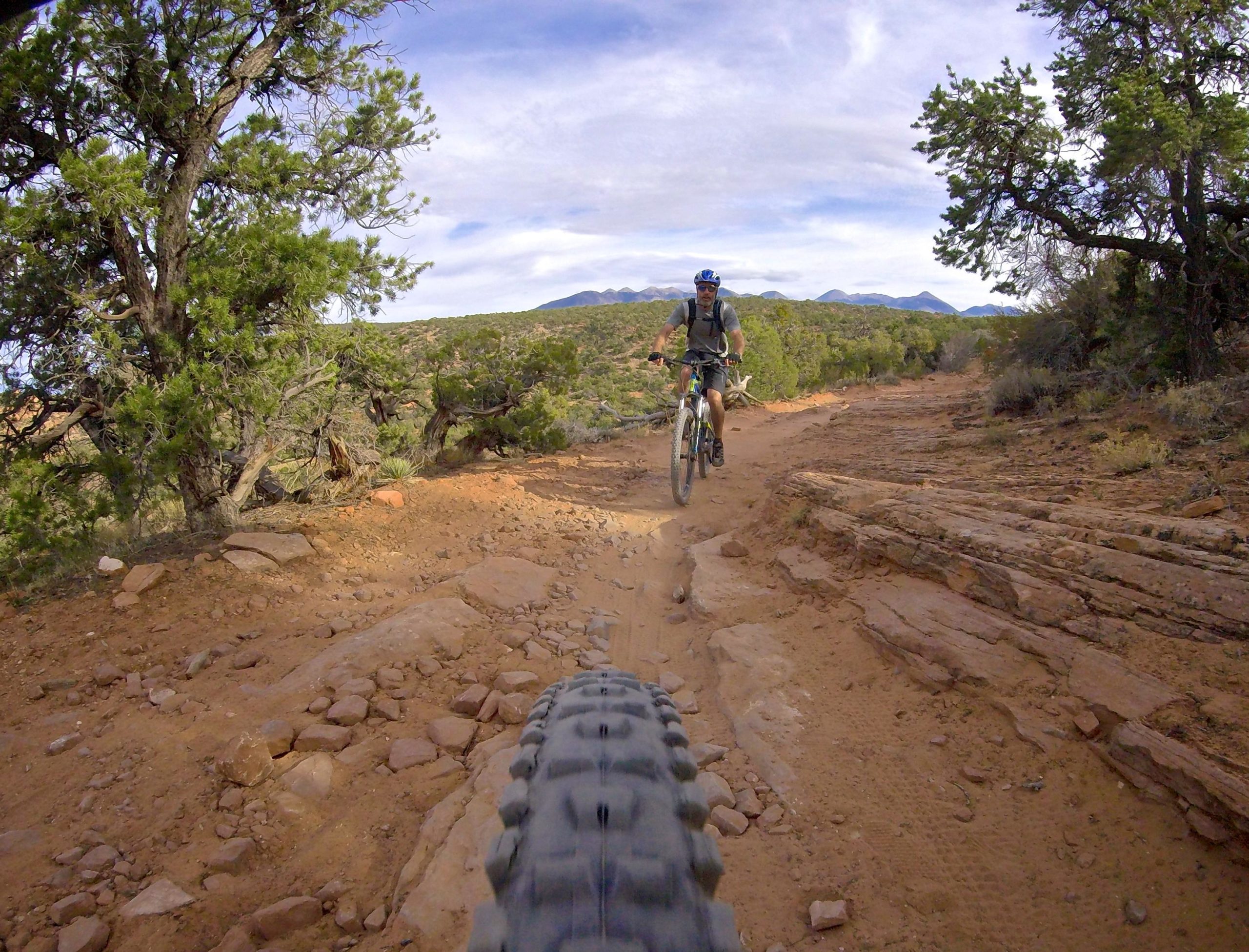 A mountain biker rides along a rocky, dirt trail surrounded by vegetation, with mountains visible in the background under a partly cloudy sky. The view is taken from the perspective of the bike, highlighting the front tire on the rugged terrain. The Whole Enchilada mountain bike trail.