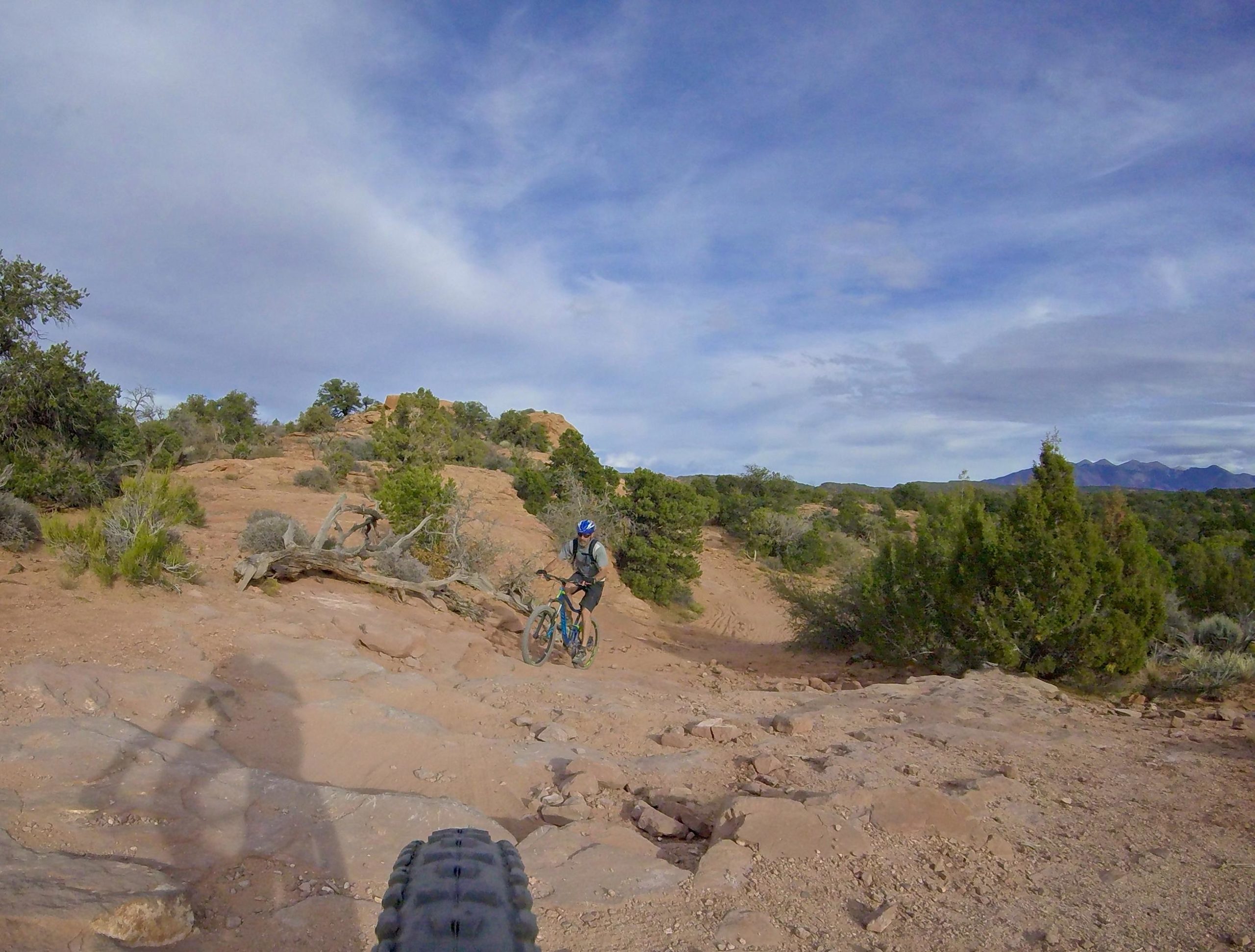 A mountain biker navigates a rocky trail in a desert landscape, surrounded by shrubs and trees, with distant mountains visible under a partly cloudy sky. The image captures the perspective from the bike's front tire. The Whole Enchilada mountain bike trail.