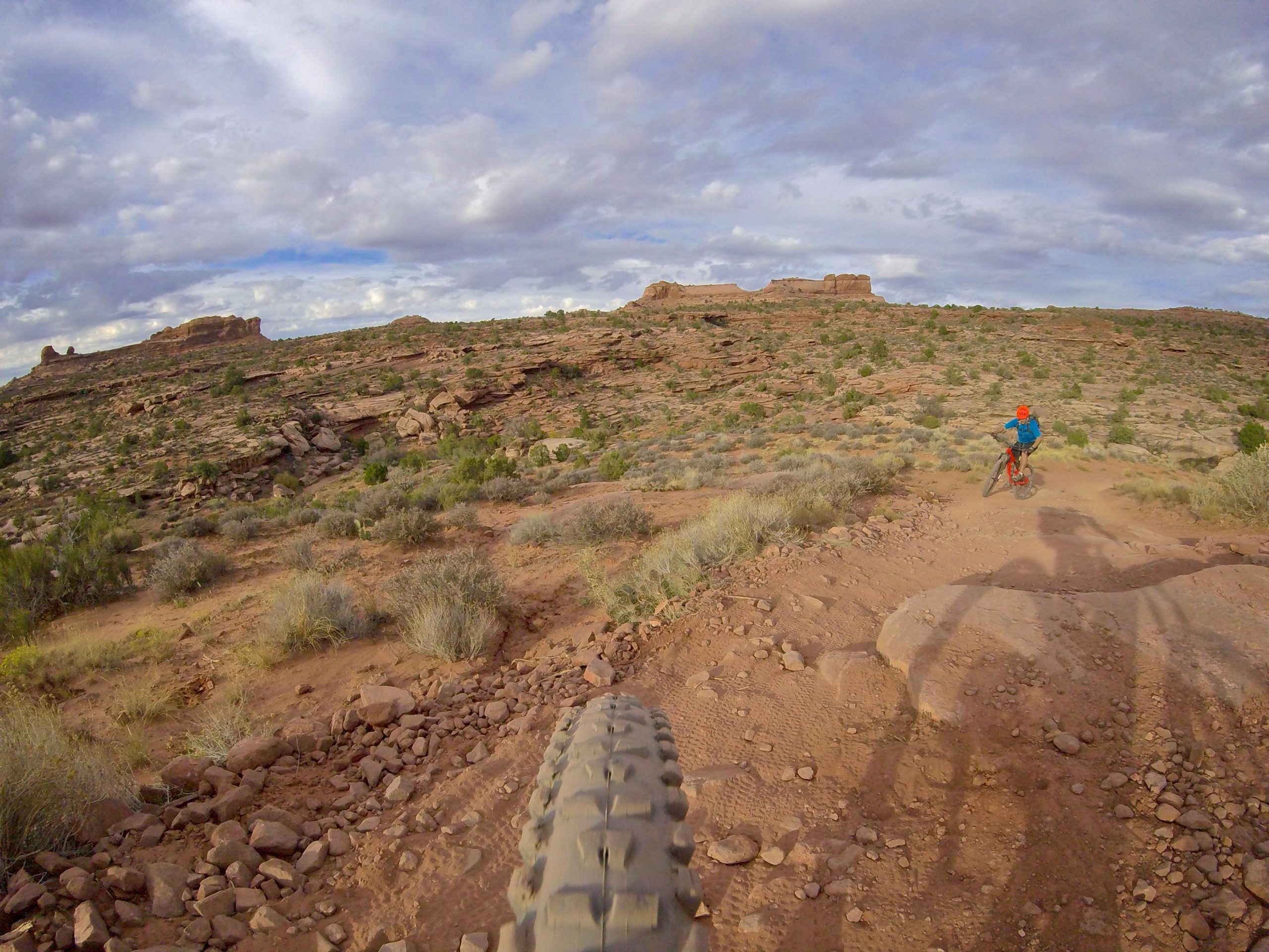 A mountain biker navigates a rocky trail in a desert landscape under a partly cloudy sky, with rugged cliffs and sparse vegetation in the background. The focus is on the bike's tire in the foreground, emphasizing the rugged terrain. The Whole Enchilada mountain bike trail.