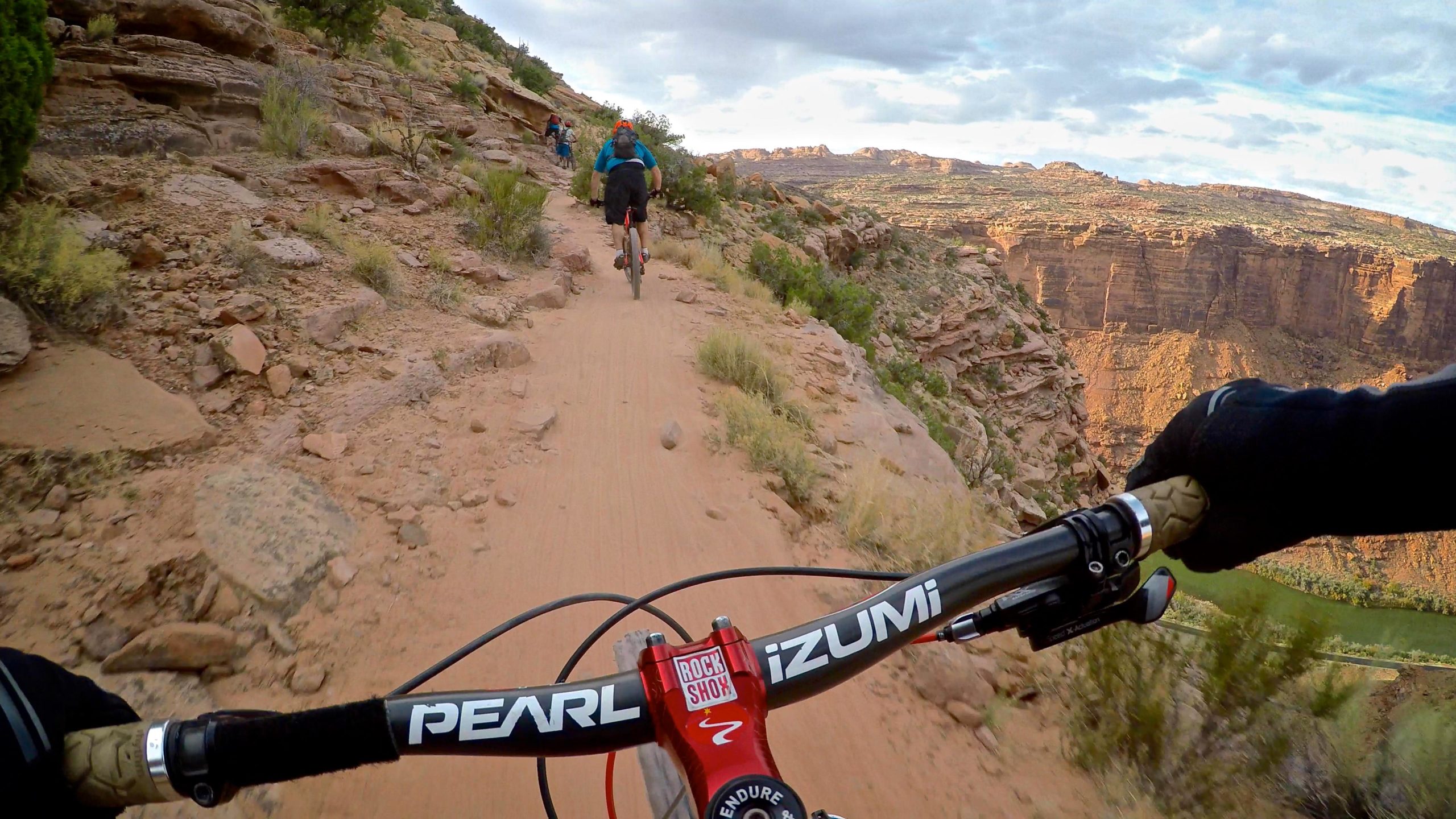 A mountain biking perspective showing the handlebars and grip of a bike on a dirt trail winding through rugged, rocky terrain. In the background, another cyclist rides ahead, with expansive views of a canyon and lush greenery below under a partly cloudy sky. The Whole Enchilada mountain bike trail.