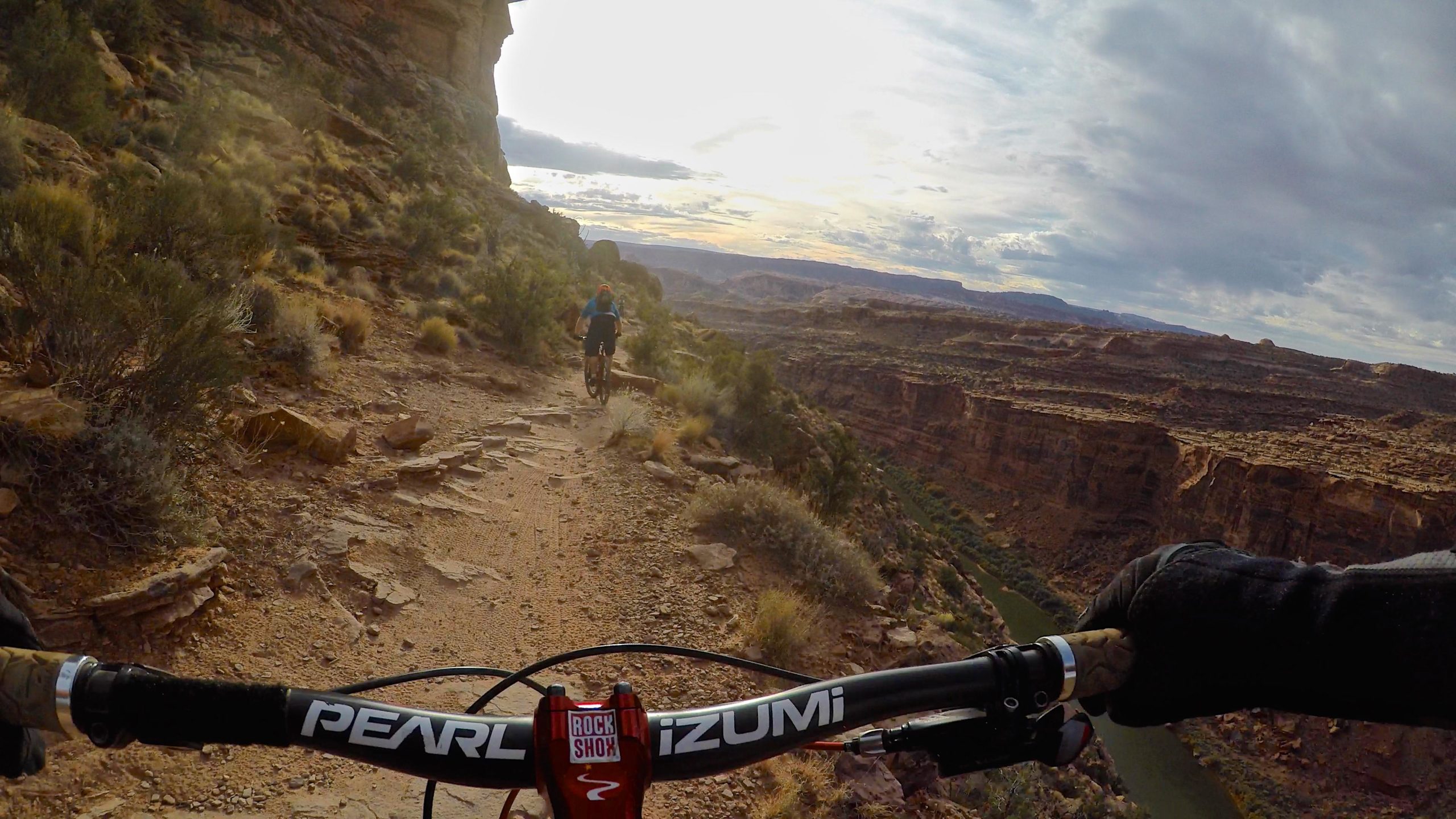 Mountain biker navigating a narrow trail along a canyon, with rocky terrain and lush vegetation visible. The background features a dramatic landscape of cliffs and a river winding through the valley under a cloudy sky. The biker is wearing a blue shirt and riding away from the camera. The Whole Enchilada mountain bike trail.