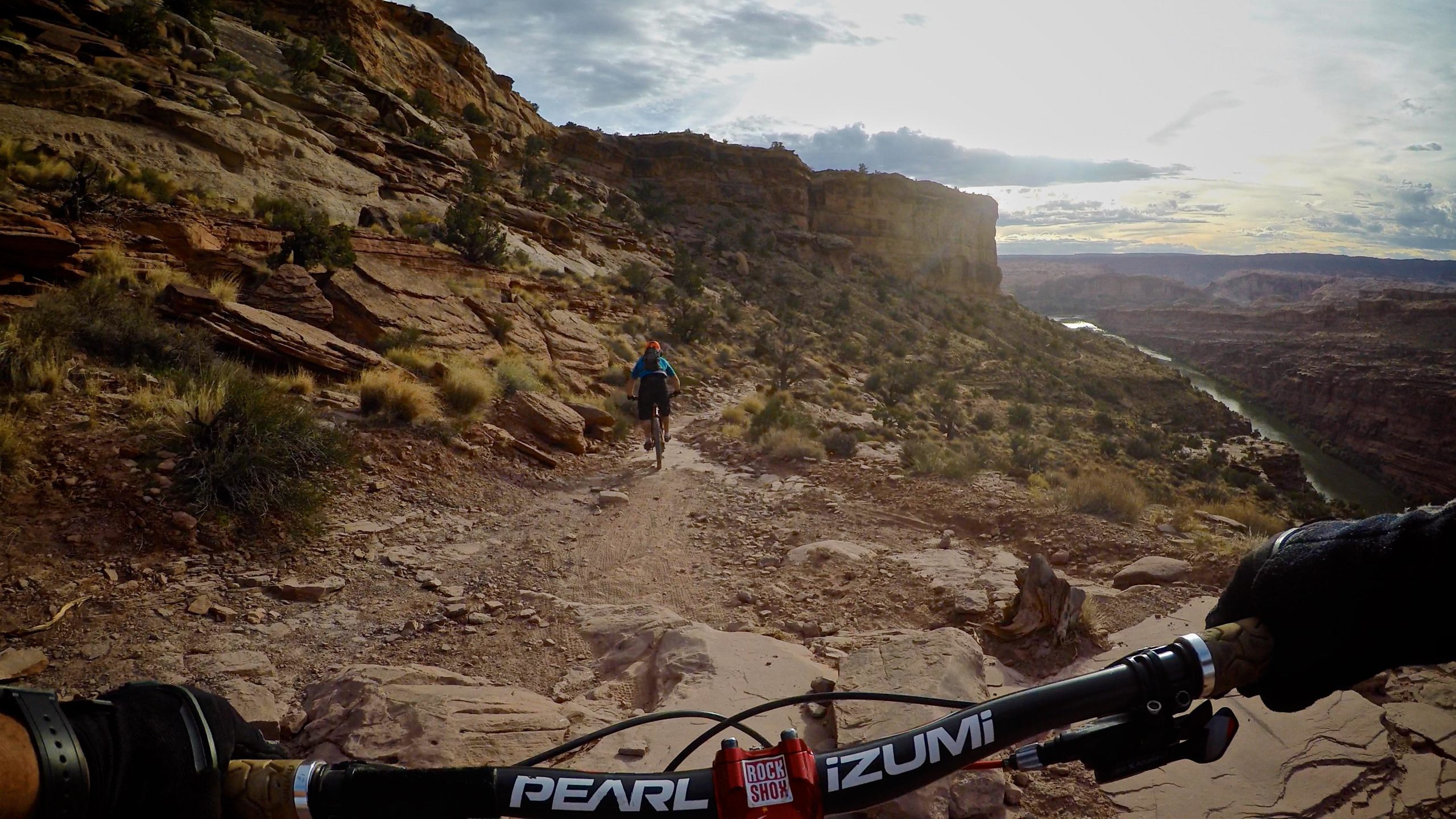 Mountain biker navigating a rocky trail with a scenic canyon view in the background. The handlebars of the bike are in the foreground, while a rider in blue can be seen pedaling ahead on the winding path. The landscape features rugged cliffs, sparse vegetation, and a river flowing through the canyon below, illuminated by soft sunlight. The Whole Enchilada mountain bike trail.