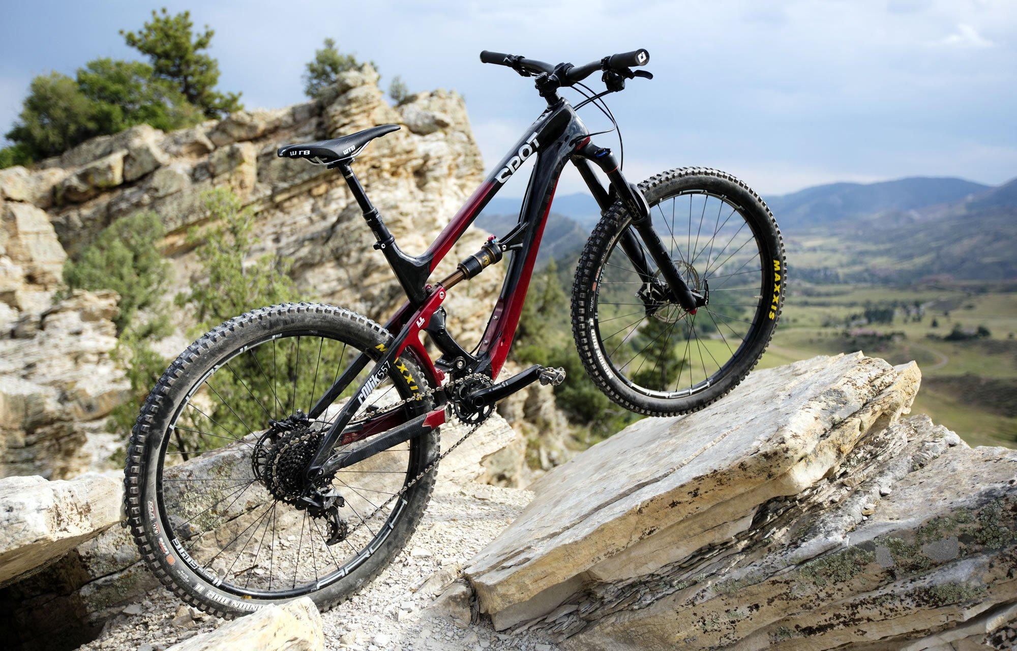 Rollik 27.5: A mountain bike perched on a rocky outcrop overlooking a scenic landscape, featuring rolling hills and distant mountains under a partly cloudy sky. The bike is primarily black with red accents and has rugged tires suitable for off-road terrain.