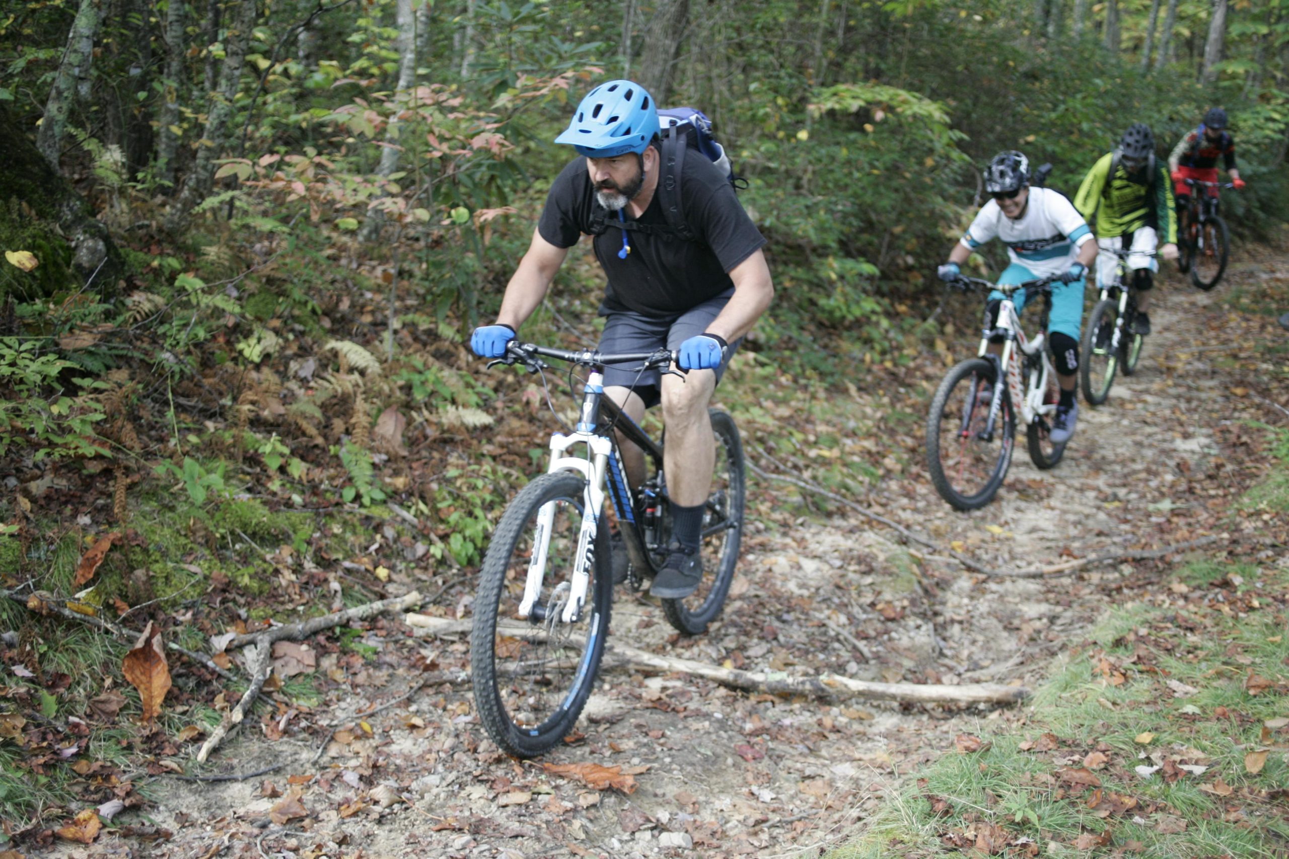 A group of four mountain bikers navigating a rugged trail surrounded by trees and autumn foliage. The leading rider is focused, wearing a blue helmet and gloves, and riding a white bicycle. The trail is rocky with scattered leaves and fallen branches. Bennett Gap / 138 mountain bike trail.