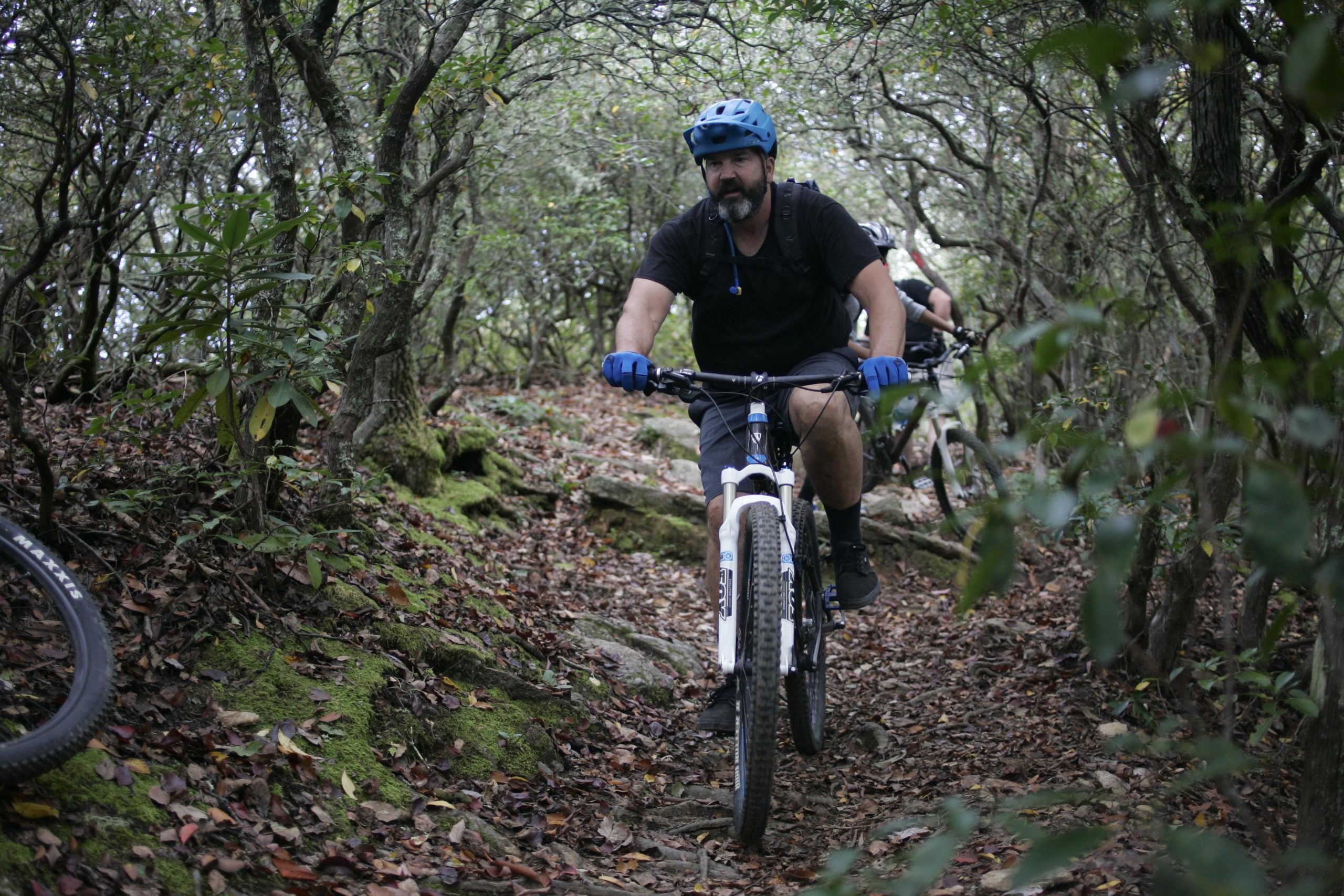 A mountain biker riding along a narrow, rocky trail in a dense forest. The biker is wearing a blue helmet and gloves, with a black shirt and shorts, navigating through the terrain surrounded by trees and fallen leaves. Another cyclist can be seen in the background. Bennett Gap / 138 mountain bike trail.