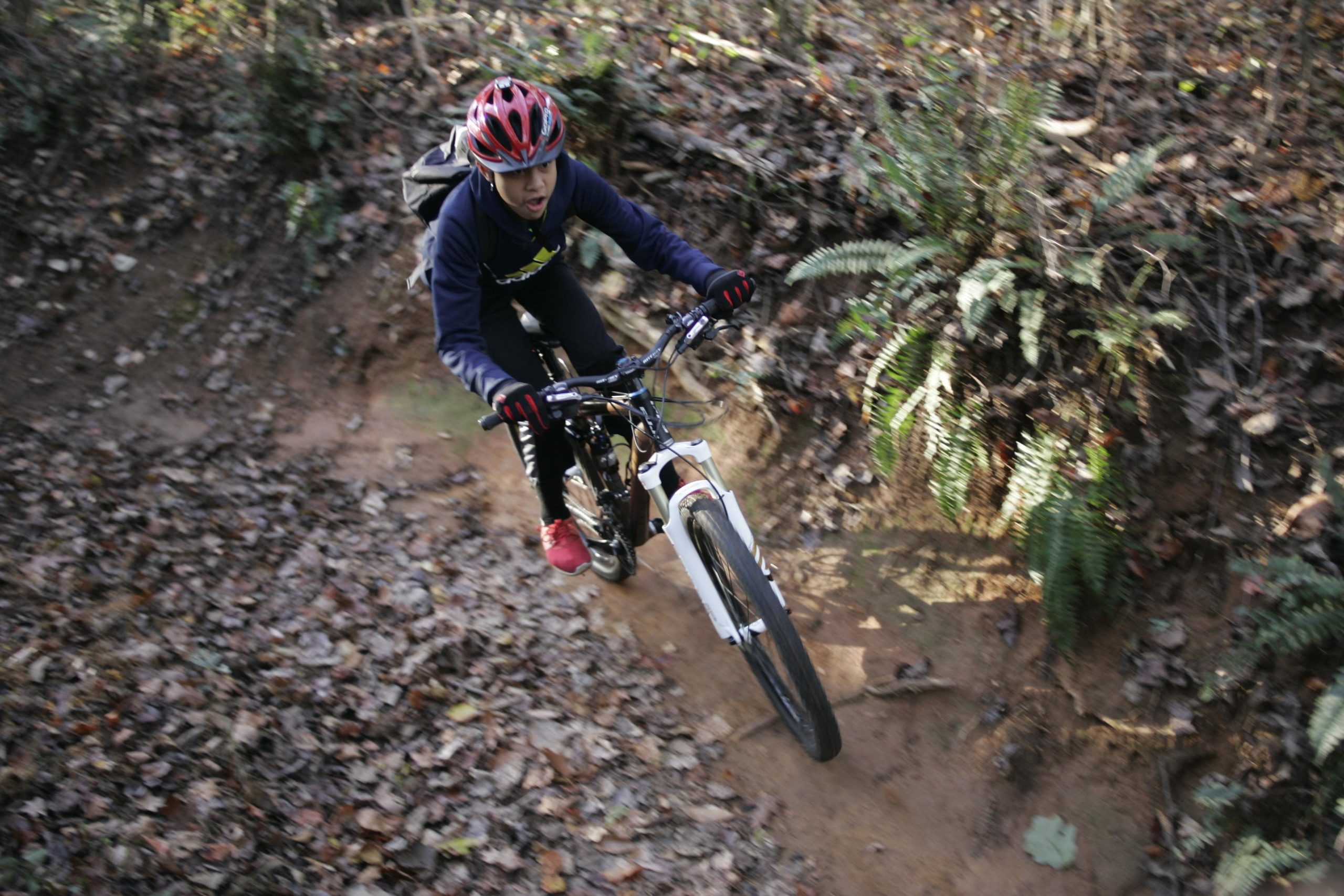 A young person riding a mountain bike on a dirt trail surrounded by fallen leaves and greenery. The rider is wearing a red helmet, gloves, and a blue sweater, focused on navigating the trail. Hobby Park mountain bike trail.