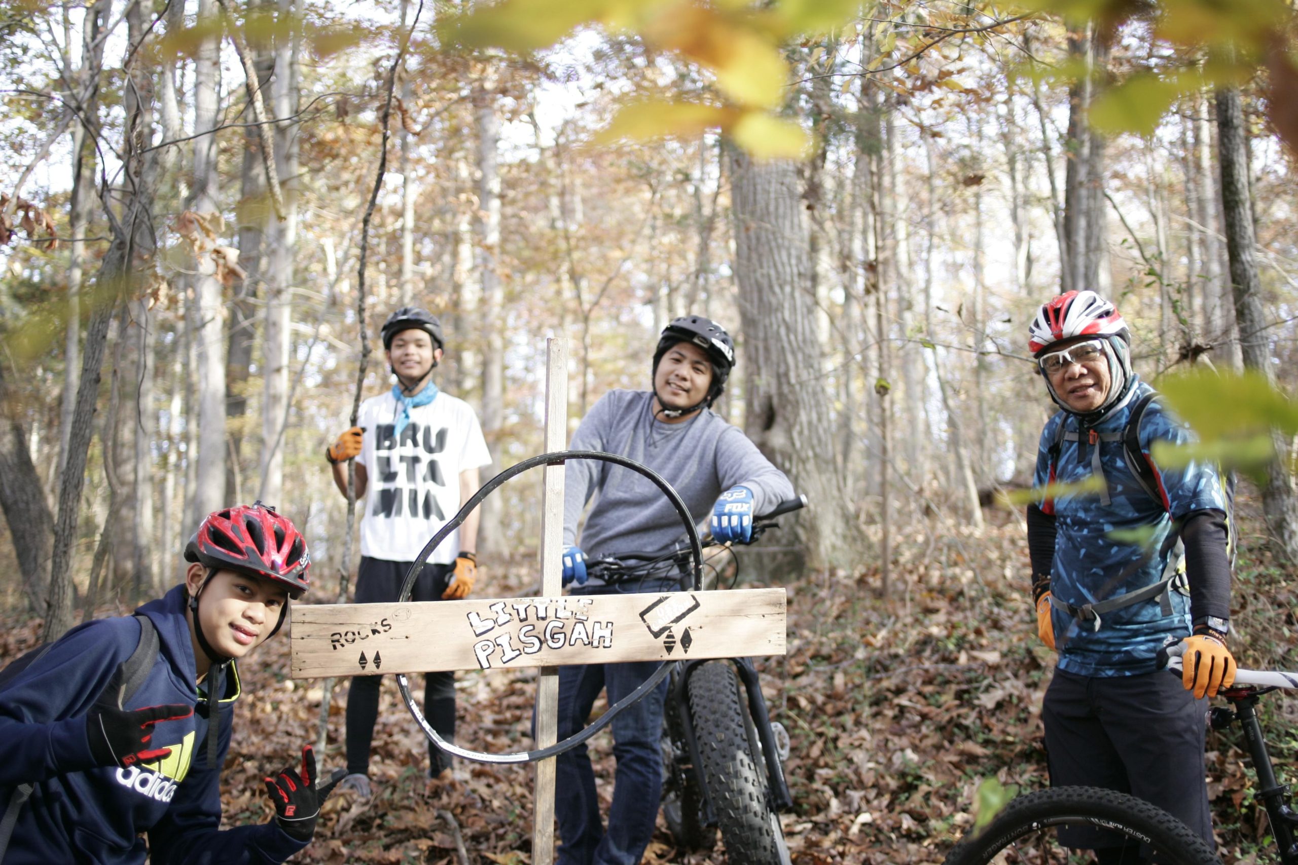 Four individuals in cycling gear pose in a wooded area, with autumn foliage in the background. They are gathered around a wooden sign labeled "Little Pisgah," and one of them is holding a bicycle. The group appears cheerful and engaged, suggesting a shared enjoyment of mountain biking. Hobby Park mountain bike trail.
