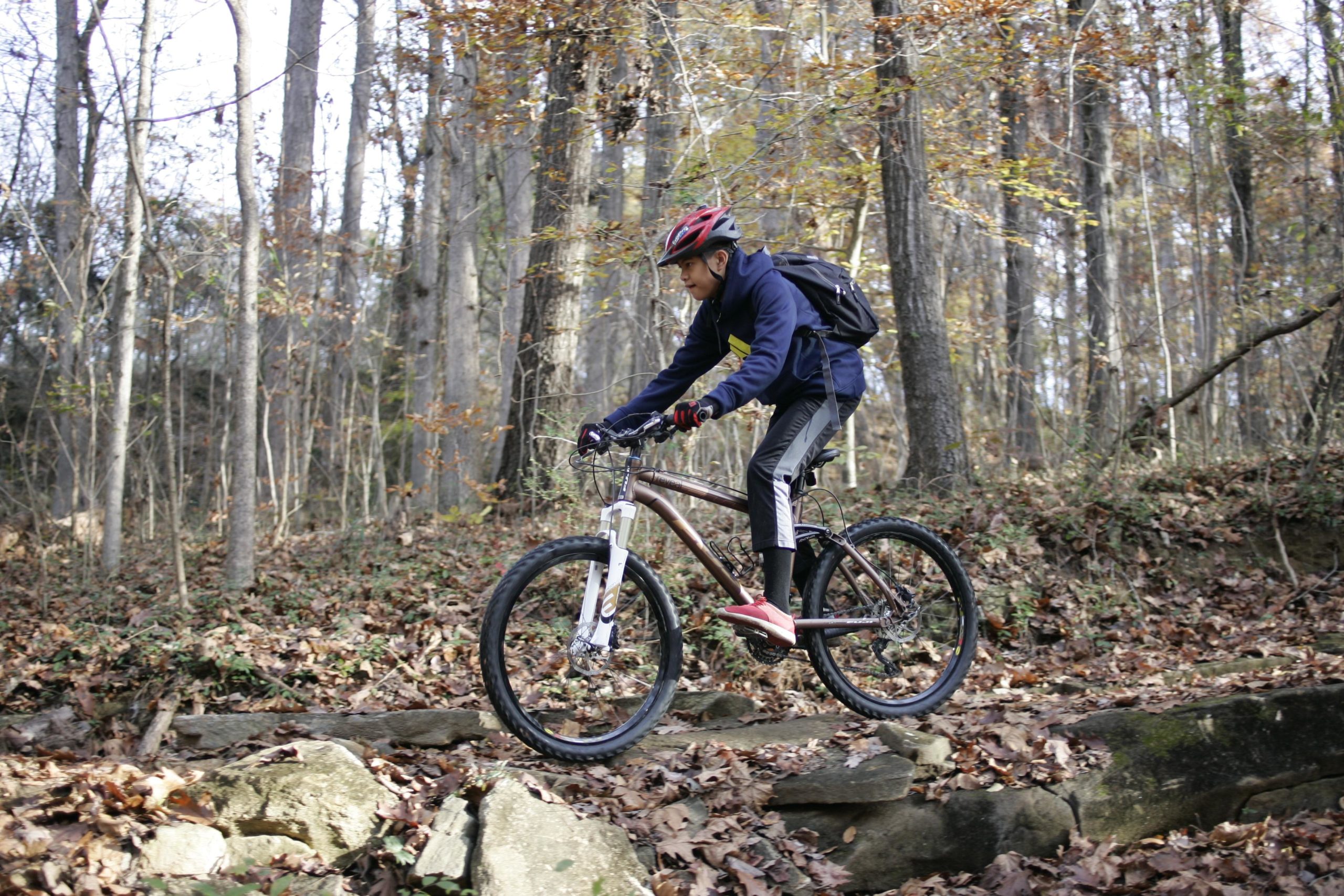 A young person riding a mountain bike over rocky terrain in a forested area, surrounded by trees with autumn foliage. The rider is wearing a helmet, a navy blue jacket, and gloves, focused on navigating the uneven ground. Hobby Park mountain bike trail.
