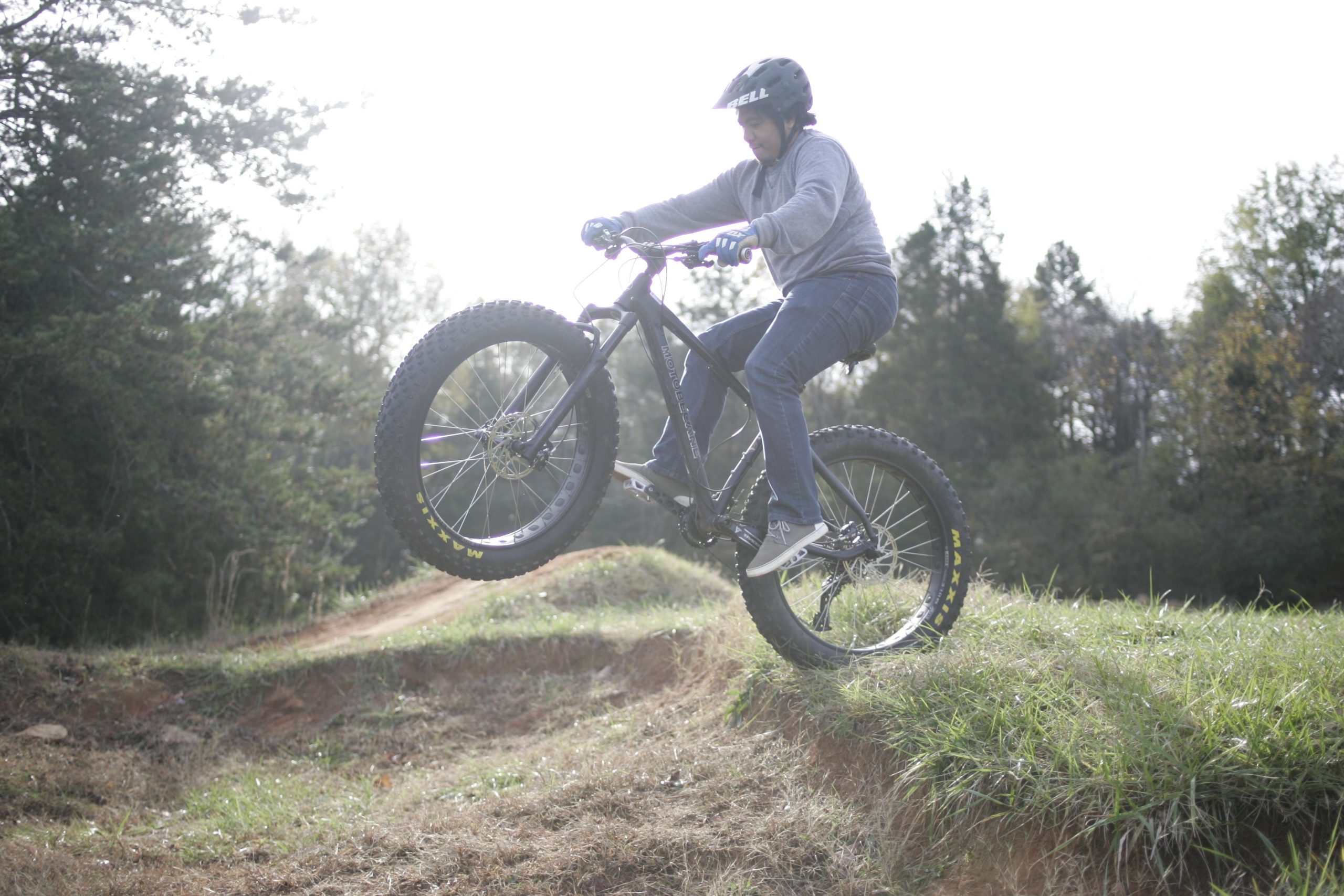 A person performing a bicycle trick by lifting the front wheel off the ground, set against a backdrop of trees and grassy terrain. The rider is wearing a helmet and gloves, and the bike features thick tires suitable for off-road riding. Hobby Park mountain bike trail.